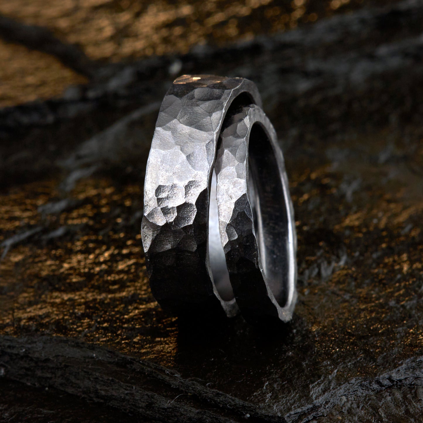 Handgefertigtes Ehering-Set aus massivem Tantal auf rustikalem Leder. Herrenring (6mm) und Damenring (4mm) mit markanter, gehämmerter Oberfläche in dunklem Anthrazit. Einzigartige Schmiedekunst für exklusive Hochzeitsringe. SEO-Keywords: Tantal Eheringe gehämmert, dunkle Trauringe, Tantalum Wedding Bands, nachhaltiger Luxusschmuck, seltene Edelmetalle Hochzeit. Sichtbare Details der individuellen Textur und des tiefen, metallischen Glanzes.