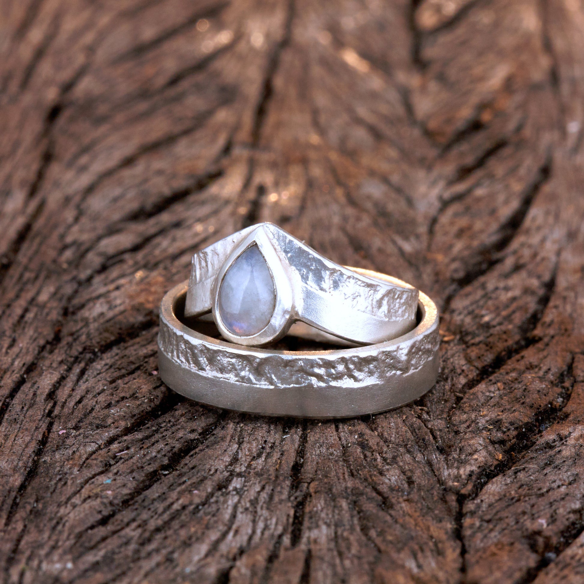 Hochwertiges Eheringe Set aus massivem 925 Sterling Silber mit kontrastreicher Oberfläche halb matt und halb retikuliert. Der Damenring in eleganter Wishbone V-Form trägt einen handgefassten Regenbogen-Mondstein im Tropfenschliff 9x6 mm mit blauem Schimmer. Der Herrenring ist 6x2 mm breit und spiegelt das maritime nordische Design wider. Handgefertigte Markenware aus der Goldschmiede Silberschweif für Paare die exklusiven Edelsteinschmuck und archaische Strukturen suchen