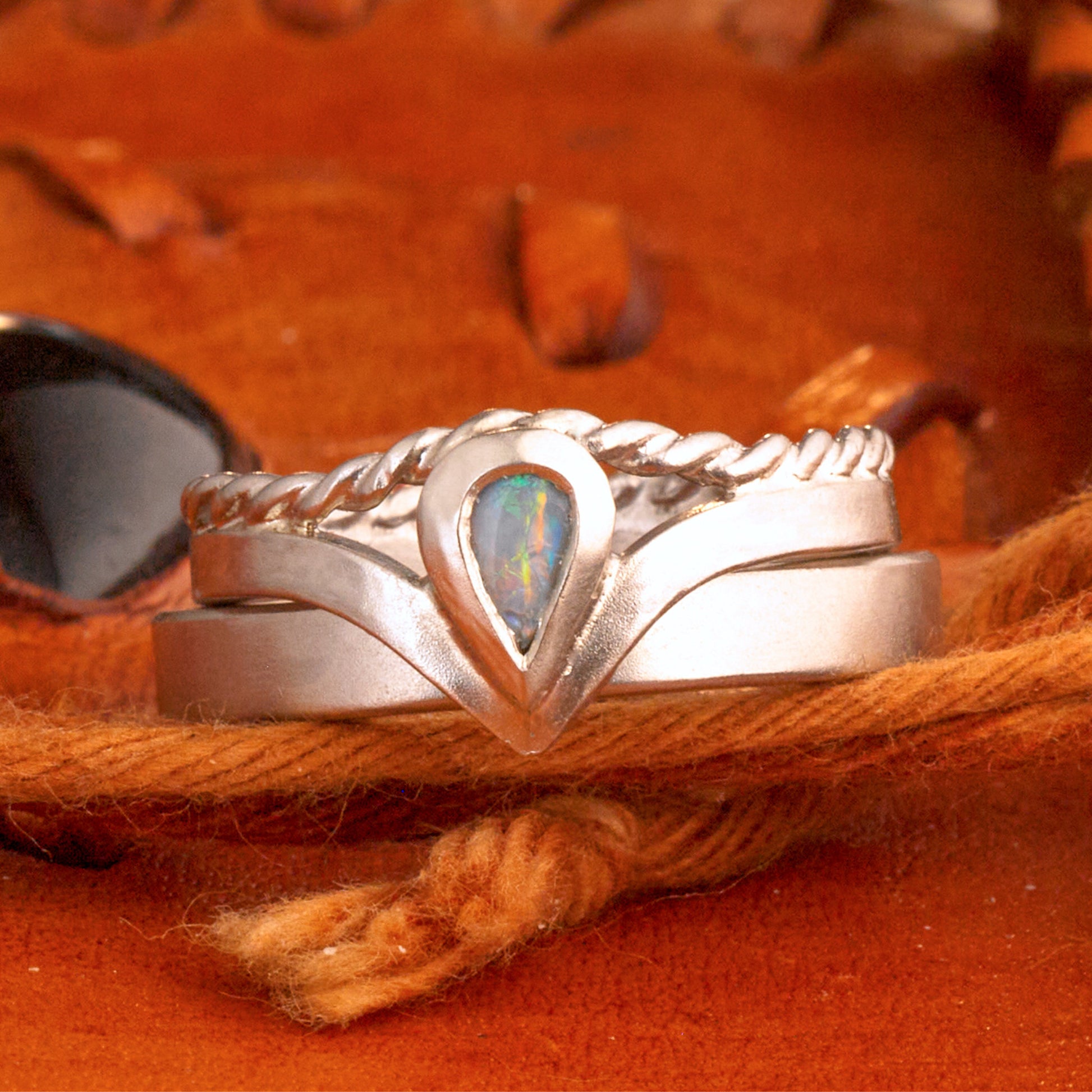 Damenring Detail: Handgefertigter Wishbone-Ring aus mattem 925 Silber von Silberschweif mit einem 6x4 mm Opaltropfen. Man erkennt das detailreiche Kordeldesign und die präzise Fassung des Edelsteins.
Set-Ansicht: Minimalistisches Trauring-Set aus Silber. Der filigrane Damenring mit Opal und Kordelschiene liegt auf dem schlichten, 4 mm breiten Herrenring vor einem rustikalen Steinhintergrund.
Herrenring Fokus: Schlichter silberner Herrenring (4x2 mm) mit matter Oberfläche, handgefertigt in Deutschland. 