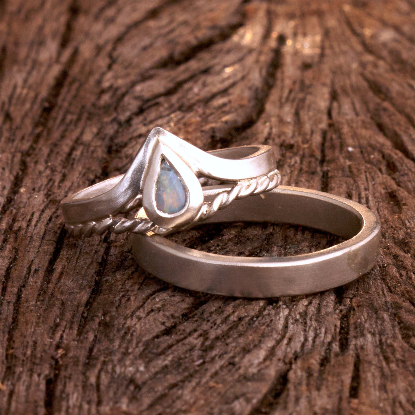 Damenring Detail: Handgefertigter Wishbone-Ring aus mattem 925 Silber von Silberschweif mit einem 6x4 mm Opaltropfen. Man erkennt das detailreiche Kordeldesign und die präzise Fassung des Edelsteins.
Set-Ansicht: Minimalistisches Trauring-Set aus Silber. Der filigrane Damenring mit Opal und Kordelschiene liegt auf dem schlichten, 4 mm breiten Herrenring vor einem rustikalen Steinhintergrund.
Herrenring Fokus: Schlichter silberner Herrenring (4x2 mm) mit matter Oberfläche, handgefertigt in Deutschland. 