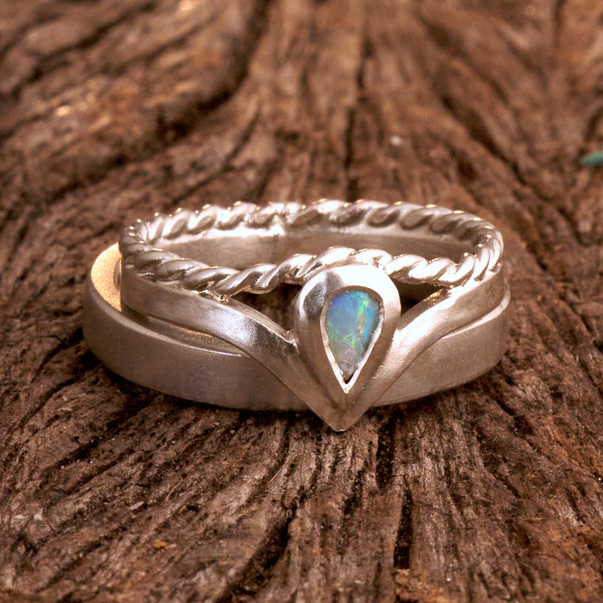 Damenring Detail: Handgefertigter Wishbone-Ring aus mattem 925 Silber von Silberschweif mit einem 6x4 mm Opaltropfen. Man erkennt das detailreiche Kordeldesign und die präzise Fassung des Edelsteins.
Set-Ansicht: Minimalistisches Trauring-Set aus Silber. Der filigrane Damenring mit Opal und Kordelschiene liegt auf dem schlichten, 4 mm breiten Herrenring vor einem rustikalen Steinhintergrund.
Herrenring Fokus: Schlichter silberner Herrenring (4x2 mm) mit matter Oberfläche, handgefertigt in Deutschland. 