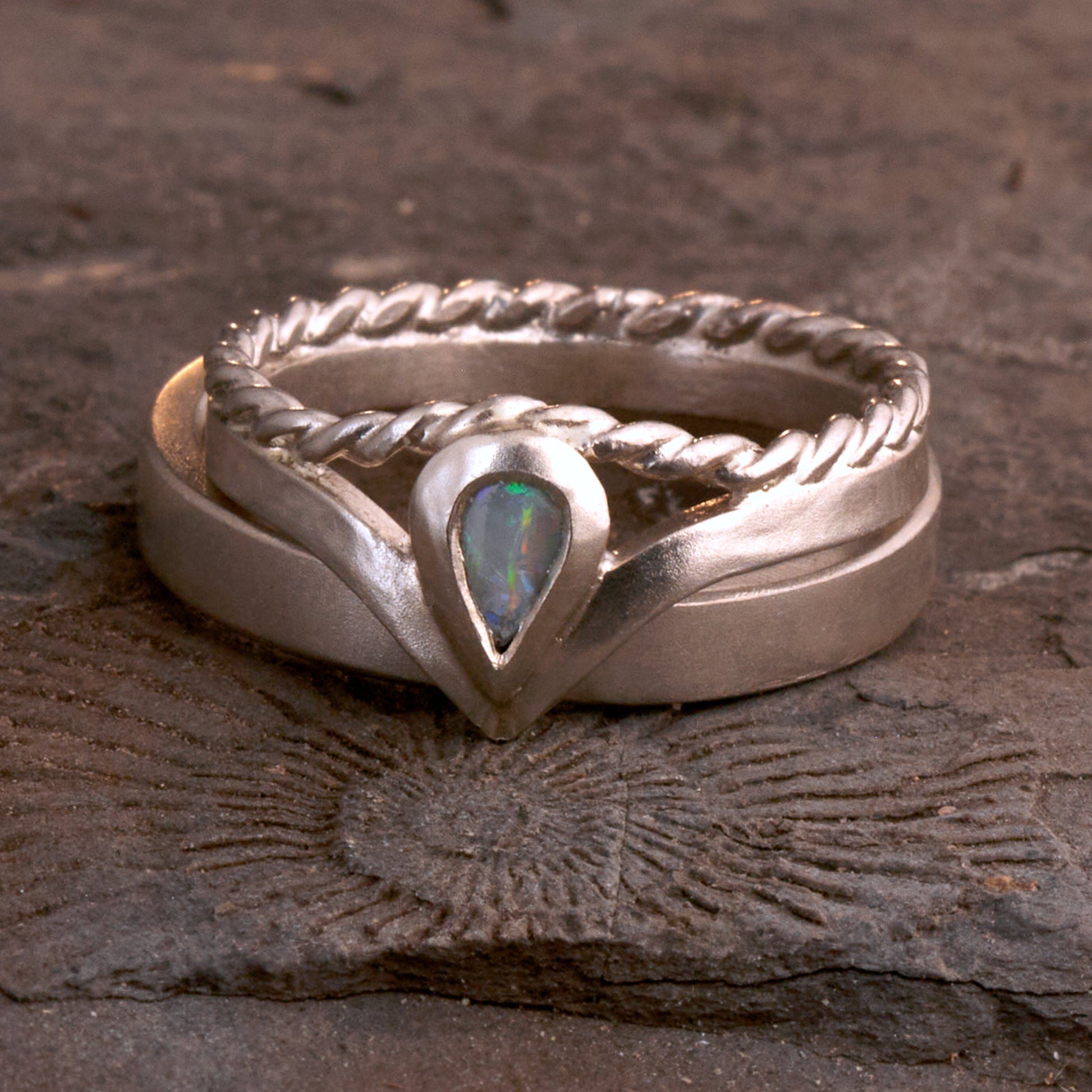 Damenring Detail: Handgefertigter Wishbone-Ring aus mattem 925 Silber von Silberschweif mit einem 6x4 mm Opaltropfen. Man erkennt das detailreiche Kordeldesign und die präzise Fassung des Edelsteins.
Set-Ansicht: Minimalistisches Trauring-Set aus Silber. Der filigrane Damenring mit Opal und Kordelschiene liegt auf dem schlichten, 4 mm breiten Herrenring vor einem rustikalen Steinhintergrund.
Herrenring Fokus: Schlichter silberner Herrenring (4x2 mm) mit matter Oberfläche, handgefertigt in Deutschland. 