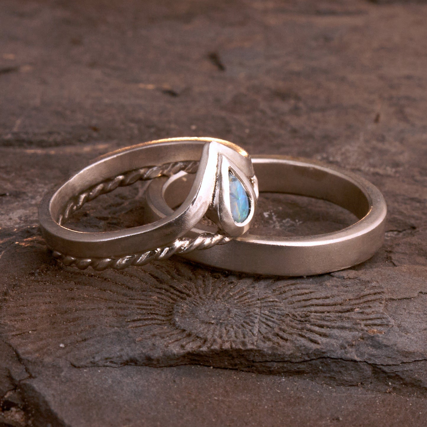 Damenring Detail: Handgefertigter Wishbone-Ring aus mattem 925 Silber von Silberschweif mit einem 6x4 mm Opaltropfen. Man erkennt das detailreiche Kordeldesign und die präzise Fassung des Edelsteins.
Set-Ansicht: Minimalistisches Trauring-Set aus Silber. Der filigrane Damenring mit Opal und Kordelschiene liegt auf dem schlichten, 4 mm breiten Herrenring vor einem rustikalen Steinhintergrund.
Herrenring Fokus: Schlichter silberner Herrenring (4x2 mm) mit matter Oberfläche, handgefertigt in Deutschland. 