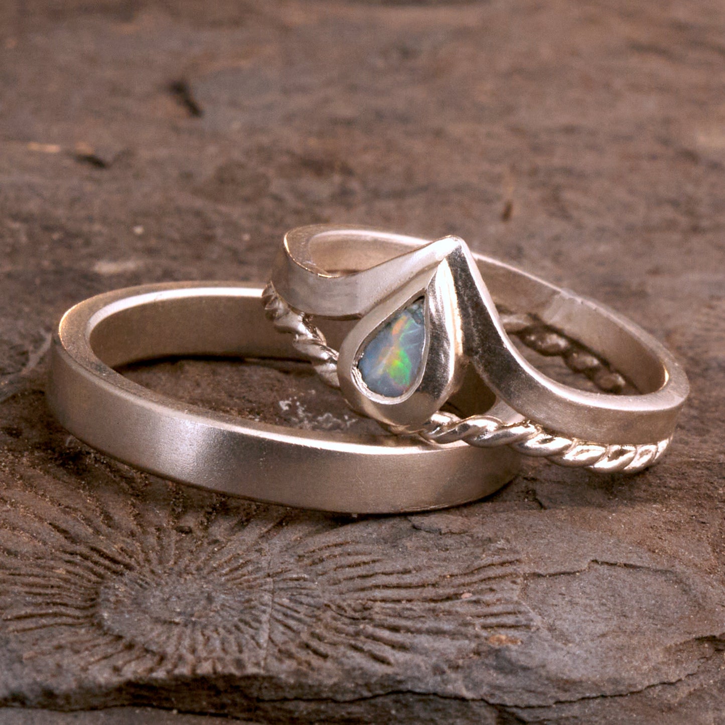 Damenring Detail: Handgefertigter Wishbone-Ring aus mattem 925 Silber von Silberschweif mit einem 6x4 mm Opaltropfen. Man erkennt das detailreiche Kordeldesign und die präzise Fassung des Edelsteins.
Set-Ansicht: Minimalistisches Trauring-Set aus Silber. Der filigrane Damenring mit Opal und Kordelschiene liegt auf dem schlichten, 4 mm breiten Herrenring vor einem rustikalen Steinhintergrund.
Herrenring Fokus: Schlichter silberner Herrenring (4x2 mm) mit matter Oberfläche, handgefertigt in Deutschland. 