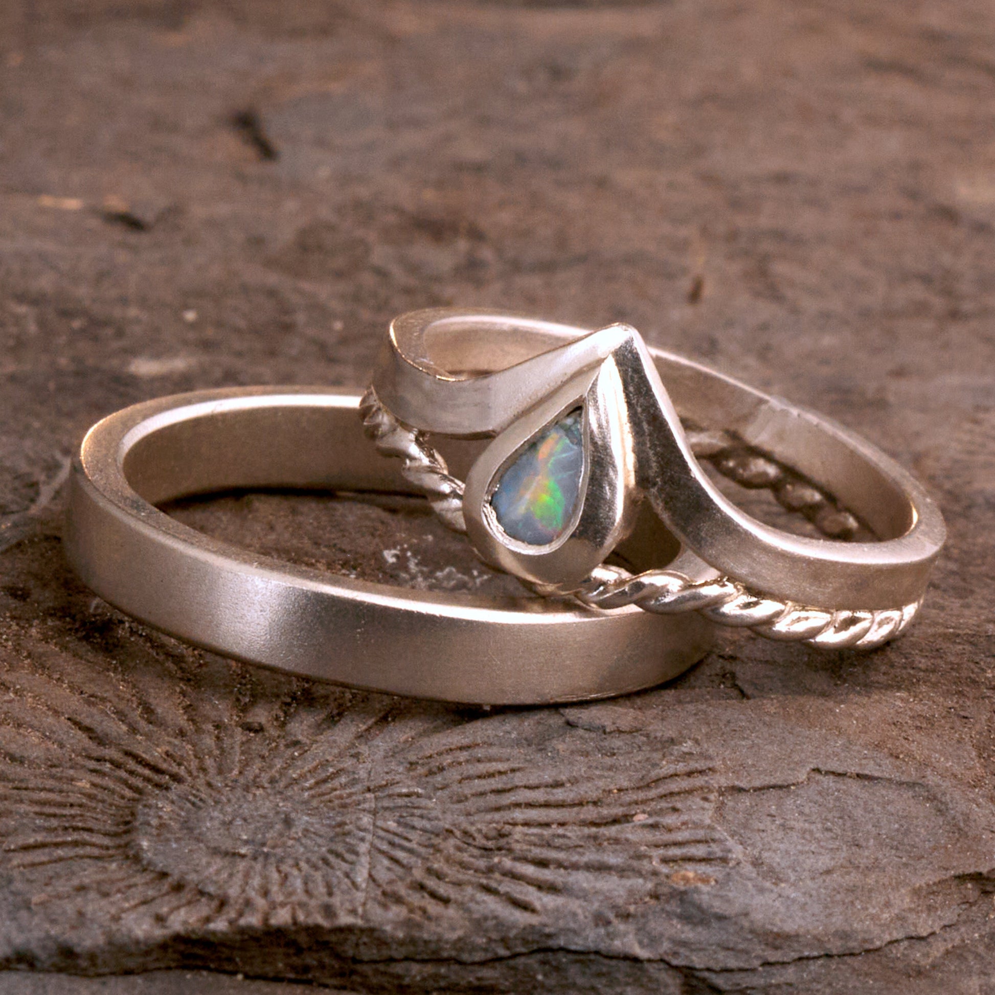 Damenring Detail: Handgefertigter Wishbone-Ring aus mattem 925 Silber von Silberschweif mit einem 6x4 mm Opaltropfen. Man erkennt das detailreiche Kordeldesign und die präzise Fassung des Edelsteins.
Set-Ansicht: Minimalistisches Trauring-Set aus Silber. Der filigrane Damenring mit Opal und Kordelschiene liegt auf dem schlichten, 4 mm breiten Herrenring vor einem rustikalen Steinhintergrund.
Herrenring Fokus: Schlichter silberner Herrenring (4x2 mm) mit matter Oberfläche, handgefertigt in Deutschland. 