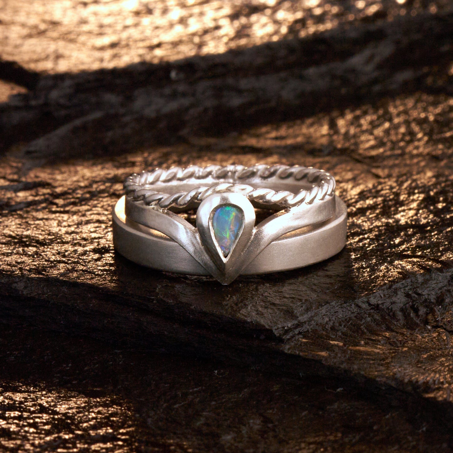 Damenring Detail: Handgefertigter Wishbone-Ring aus mattem 925 Silber von Silberschweif mit einem 6x4 mm Opaltropfen. Man erkennt das detailreiche Kordeldesign und die präzise Fassung des Edelsteins.
Set-Ansicht: Minimalistisches Trauring-Set aus Silber. Der filigrane Damenring mit Opal und Kordelschiene liegt auf dem schlichten, 4 mm breiten Herrenring vor einem rustikalen Steinhintergrund.
Herrenring Fokus: Schlichter silberner Herrenring (4x2 mm) mit matter Oberfläche, handgefertigt in Deutschland. 