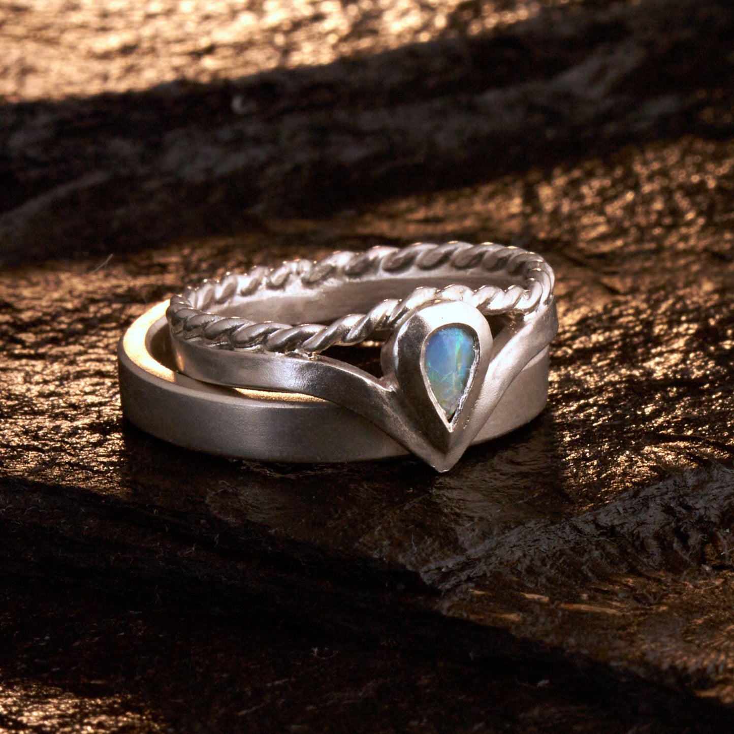 Damenring Detail: Handgefertigter Wishbone-Ring aus mattem 925 Silber von Silberschweif mit einem 6x4 mm Opaltropfen. Man erkennt das detailreiche Kordeldesign und die präzise Fassung des Edelsteins.
Set-Ansicht: Minimalistisches Trauring-Set aus Silber. Der filigrane Damenring mit Opal und Kordelschiene liegt auf dem schlichten, 4 mm breiten Herrenring vor einem rustikalen Steinhintergrund.
Herrenring Fokus: Schlichter silberner Herrenring (4x2 mm) mit matter Oberfläche, handgefertigt in Deutschland. 