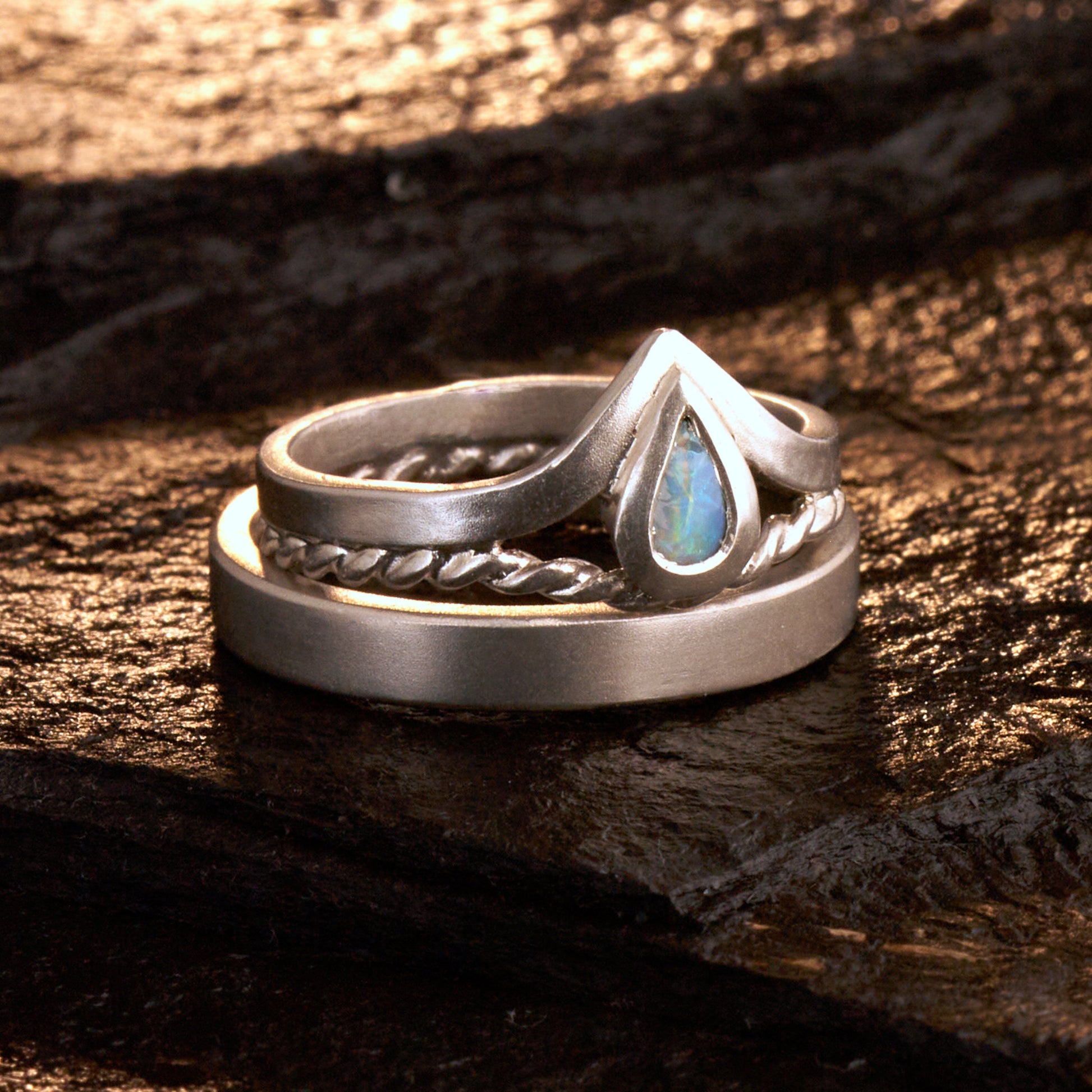 Damenring Detail: Handgefertigter Wishbone-Ring aus mattem 925 Silber von Silberschweif mit einem 6x4 mm Opaltropfen. Man erkennt das detailreiche Kordeldesign und die präzise Fassung des Edelsteins.
Set-Ansicht: Minimalistisches Trauring-Set aus Silber. Der filigrane Damenring mit Opal und Kordelschiene liegt auf dem schlichten, 4 mm breiten Herrenring vor einem rustikalen Steinhintergrund.
Herrenring Fokus: Schlichter silberner Herrenring (4x2 mm) mit matter Oberfläche, handgefertigt in Deutschland. 