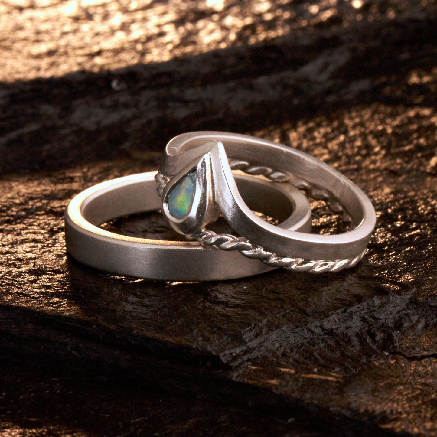 Damenring Detail: Handgefertigter Wishbone-Ring aus mattem 925 Silber von Silberschweif mit einem 6x4 mm Opaltropfen. Man erkennt das detailreiche Kordeldesign und die präzise Fassung des Edelsteins.
Set-Ansicht: Minimalistisches Trauring-Set aus Silber. Der filigrane Damenring mit Opal und Kordelschiene liegt auf dem schlichten, 4 mm breiten Herrenring vor einem rustikalen Steinhintergrund.
Herrenring Fokus: Schlichter silberner Herrenring (4x2 mm) mit matter Oberfläche, handgefertigt in Deutschland. 