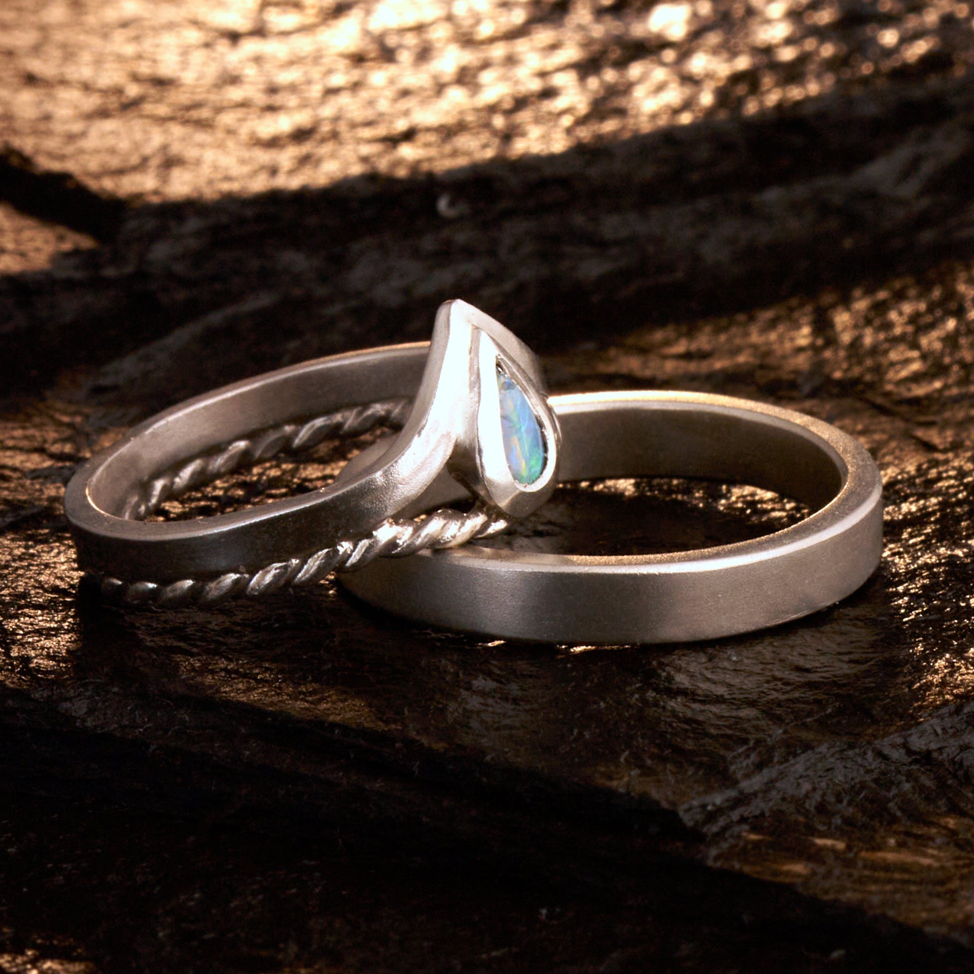 Damenring Detail: Handgefertigter Wishbone-Ring aus mattem 925 Silber von Silberschweif mit einem 6x4 mm Opaltropfen. Man erkennt das detailreiche Kordeldesign und die präzise Fassung des Edelsteins.
Set-Ansicht: Minimalistisches Trauring-Set aus Silber. Der filigrane Damenring mit Opal und Kordelschiene liegt auf dem schlichten, 4 mm breiten Herrenring vor einem rustikalen Steinhintergrund.
Herrenring Fokus: Schlichter silberner Herrenring (4x2 mm) mit matter Oberfläche, handgefertigt in Deutschland. 