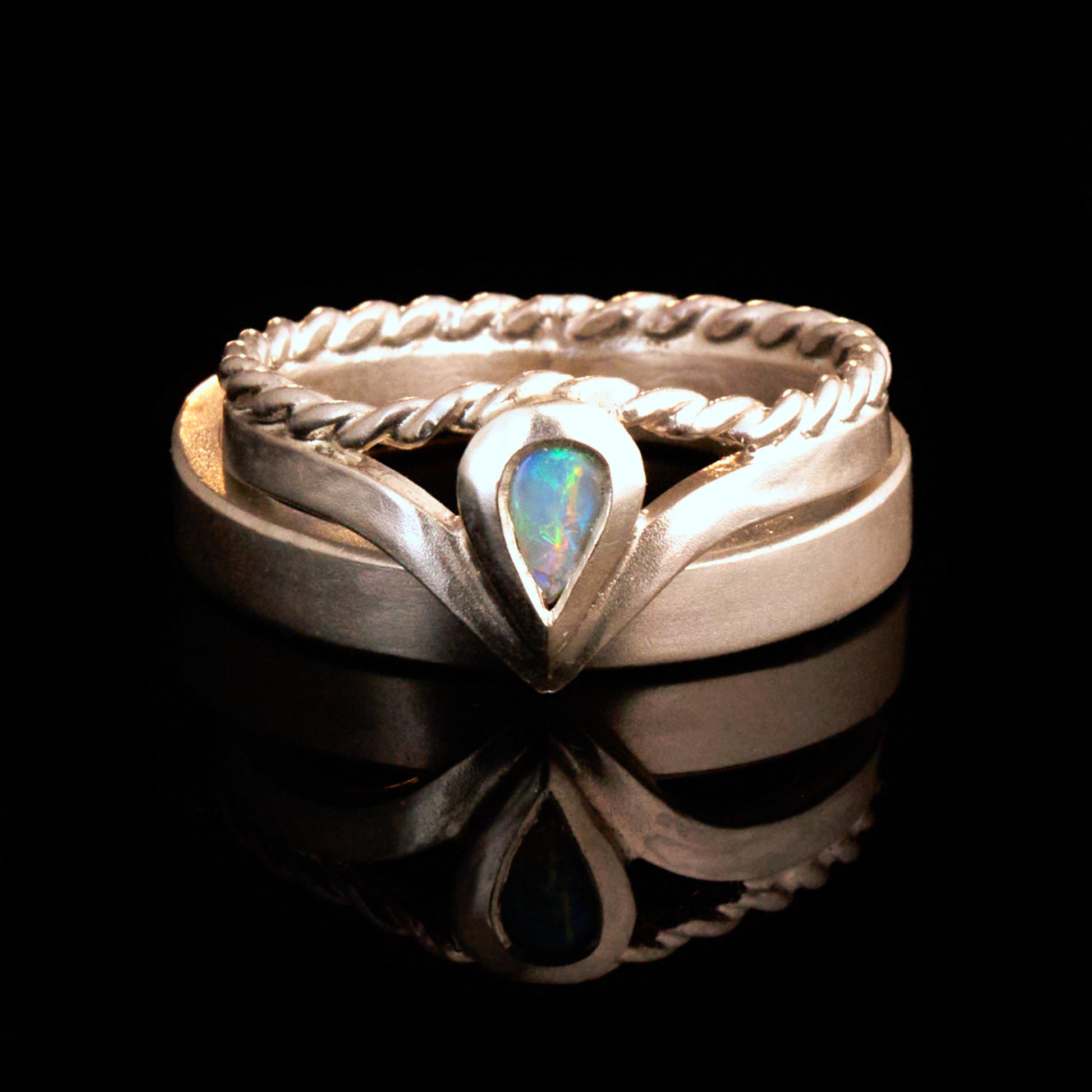 Damenring Detail: Handgefertigter Wishbone-Ring aus mattem 925 Silber von Silberschweif mit einem 6x4 mm Opaltropfen. Man erkennt das detailreiche Kordeldesign und die präzise Fassung des Edelsteins.
Set-Ansicht: Minimalistisches Trauring-Set aus Silber. Der filigrane Damenring mit Opal und Kordelschiene liegt auf dem schlichten, 4 mm breiten Herrenring vor einem rustikalen Steinhintergrund.
Herrenring Fokus: Schlichter silberner Herrenring (4x2 mm) mit matter Oberfläche, handgefertigt in Deutschland. 