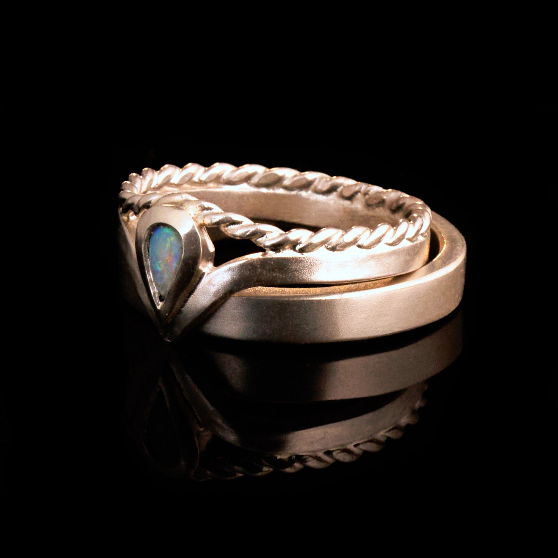 Damenring Detail: Handgefertigter Wishbone-Ring aus mattem 925 Silber von Silberschweif mit einem 6x4 mm Opaltropfen. Man erkennt das detailreiche Kordeldesign und die präzise Fassung des Edelsteins.
Set-Ansicht: Minimalistisches Trauring-Set aus Silber. Der filigrane Damenring mit Opal und Kordelschiene liegt auf dem schlichten, 4 mm breiten Herrenring vor einem rustikalen Steinhintergrund.
Herrenring Fokus: Schlichter silberner Herrenring (4x2 mm) mit matter Oberfläche, handgefertigt in Deutschland. 