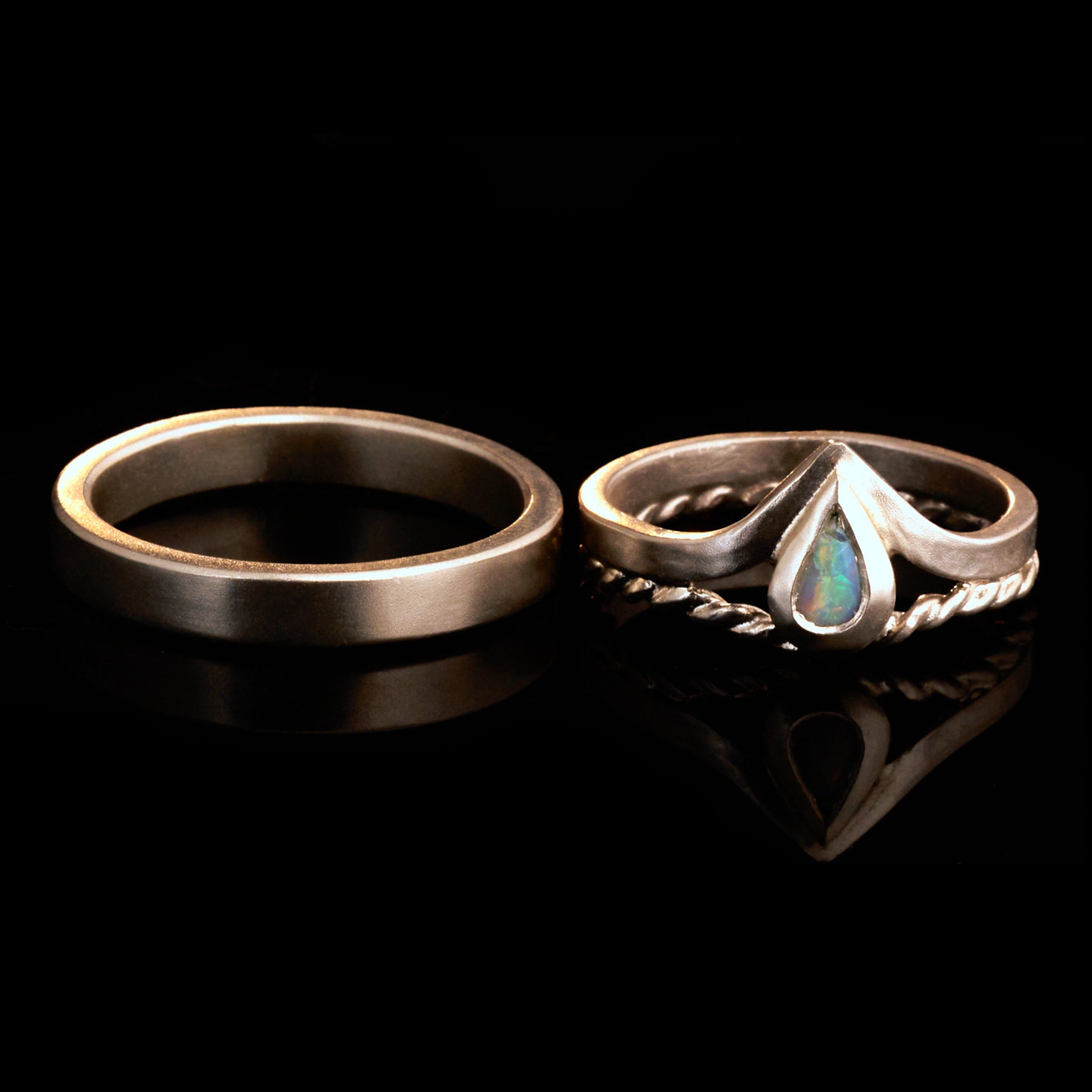 Damenring Detail: Handgefertigter Wishbone-Ring aus mattem 925 Silber von Silberschweif mit einem 6x4 mm Opaltropfen. Man erkennt das detailreiche Kordeldesign und die präzise Fassung des Edelsteins.
Set-Ansicht: Minimalistisches Trauring-Set aus Silber. Der filigrane Damenring mit Opal und Kordelschiene liegt auf dem schlichten, 4 mm breiten Herrenring vor einem rustikalen Steinhintergrund.
Herrenring Fokus: Schlichter silberner Herrenring (4x2 mm) mit matter Oberfläche, handgefertigt in Deutschland. 