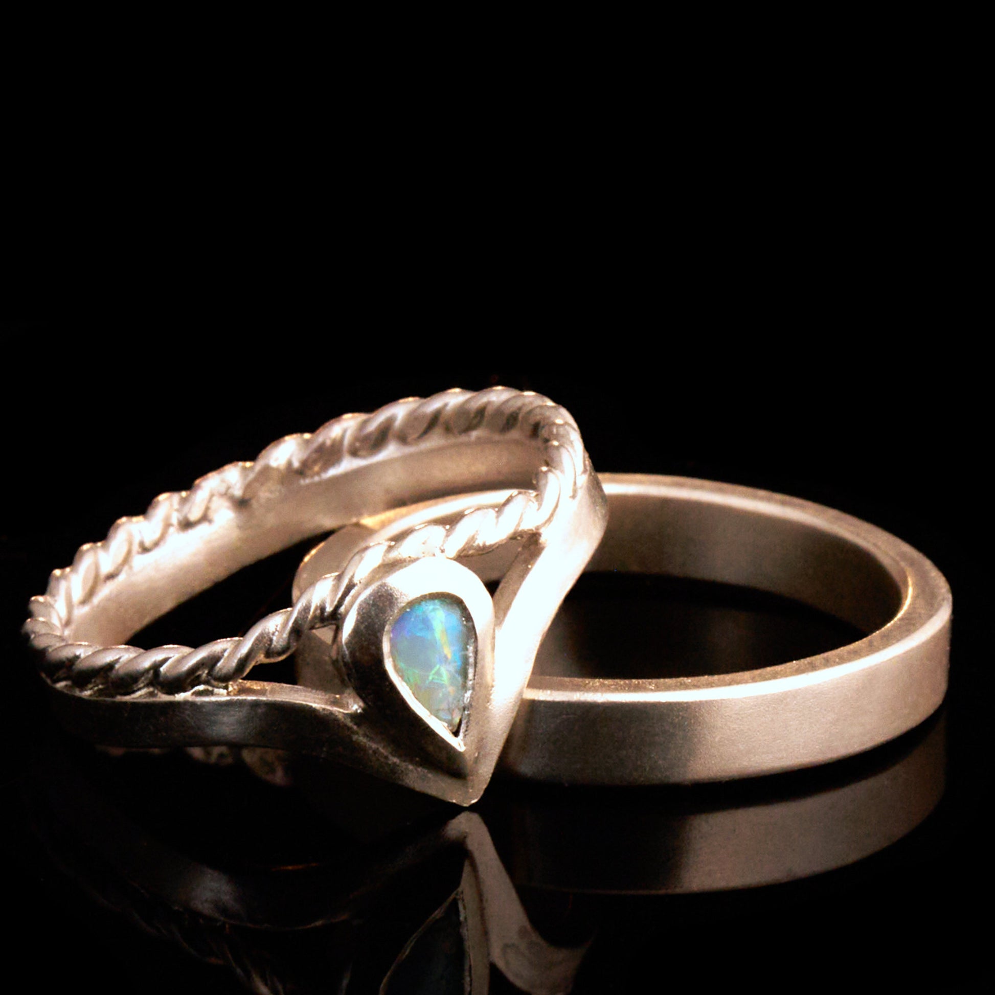 Damenring Detail: Handgefertigter Wishbone-Ring aus mattem 925 Silber von Silberschweif mit einem 6x4 mm Opaltropfen. Man erkennt das detailreiche Kordeldesign und die präzise Fassung des Edelsteins.
Set-Ansicht: Minimalistisches Trauring-Set aus Silber. Der filigrane Damenring mit Opal und Kordelschiene liegt auf dem schlichten, 4 mm breiten Herrenring vor einem rustikalen Steinhintergrund.
Herrenring Fokus: Schlichter silberner Herrenring (4x2 mm) mit matter Oberfläche, handgefertigt in Deutschland. 