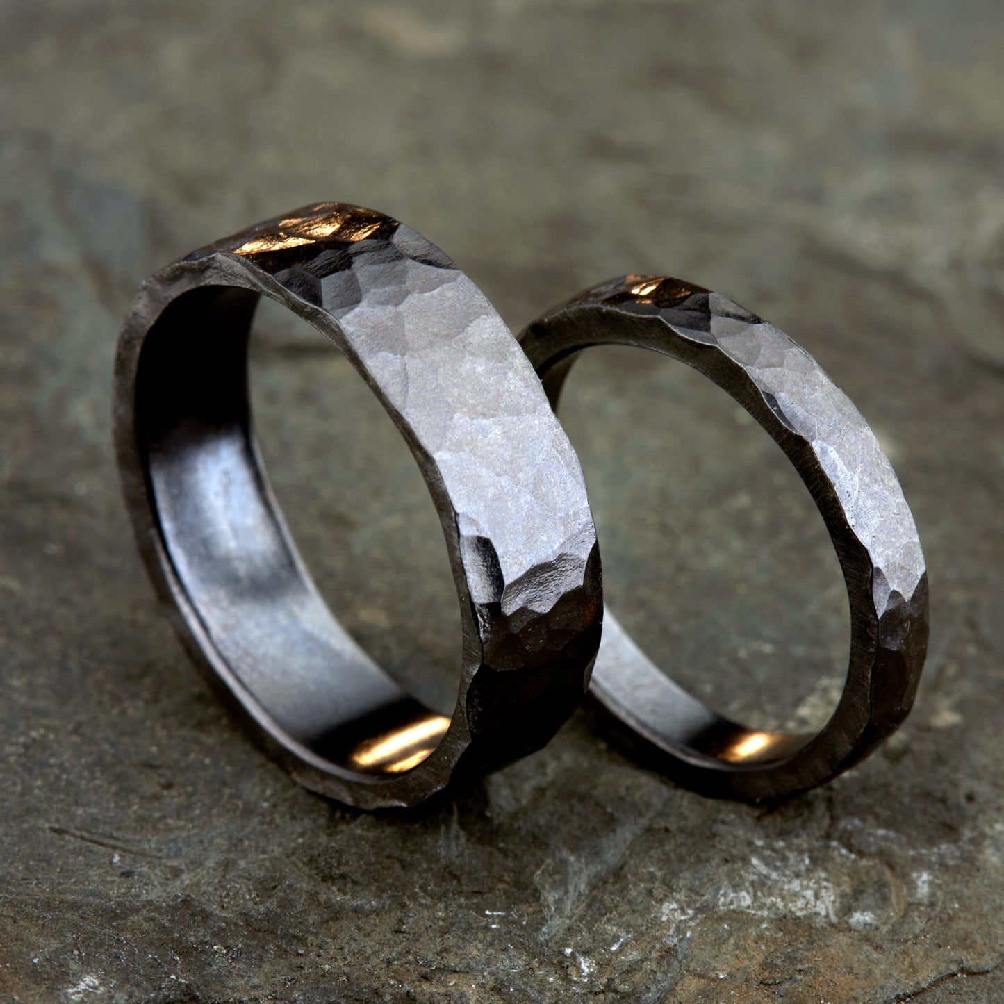 Handgefertigtes Ehering-Set aus massivem Tantal auf rustikalem Leder. Herrenring (6mm) und Damenring (4mm) mit markanter, gehämmerter Oberfläche in dunklem Anthrazit. Einzigartige Schmiedekunst für exklusive Hochzeitsringe. SEO-Keywords: Tantal Eheringe gehämmert, dunkle Trauringe, Tantalum Wedding Bands, nachhaltiger Luxusschmuck, seltene Edelmetalle Hochzeit. Sichtbare Details der individuellen Textur und des tiefen, metallischen Glanzes.
