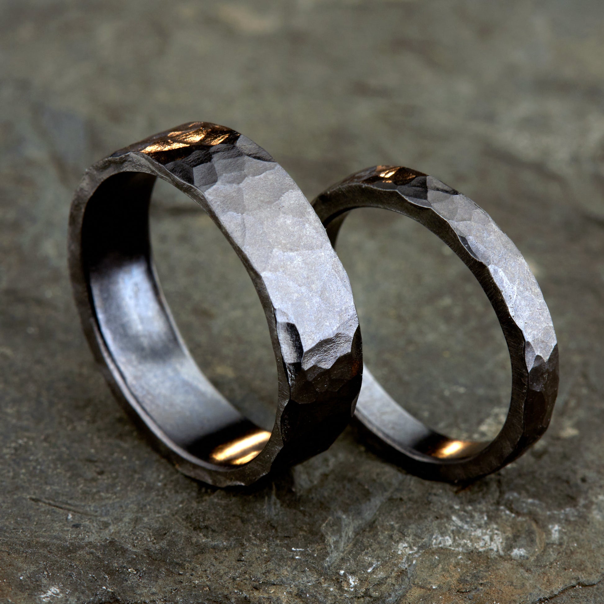 Handgefertigtes Ehering-Set aus massivem Tantal auf rustikalem Leder. Herrenring (6mm) und Damenring (4mm) mit markanter, gehämmerter Oberfläche in dunklem Anthrazit. Einzigartige Schmiedekunst für exklusive Hochzeitsringe. SEO-Keywords: Tantal Eheringe gehämmert, dunkle Trauringe, Tantalum Wedding Bands, nachhaltiger Luxusschmuck, seltene Edelmetalle Hochzeit. Sichtbare Details der individuellen Textur und des tiefen, metallischen Glanzes.
