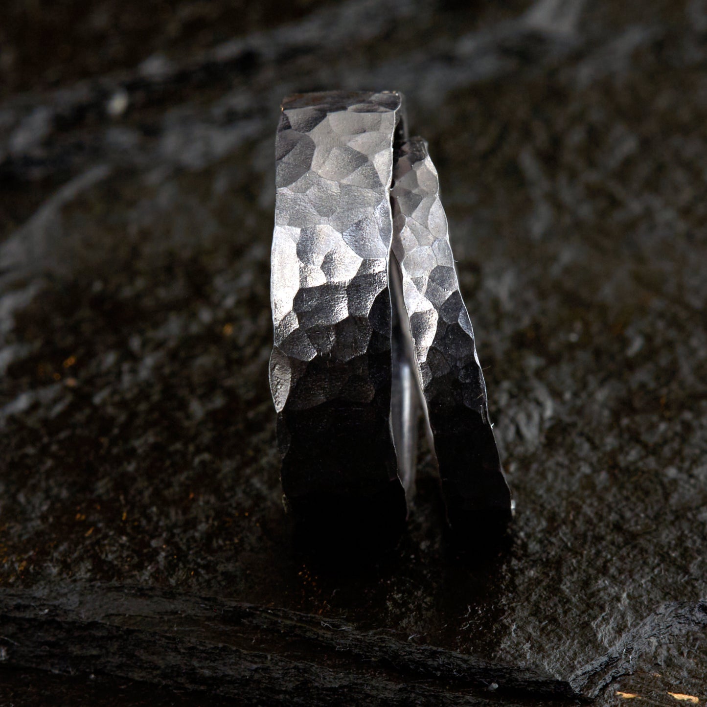 Handgefertigtes Ehering-Set aus massivem Tantal auf rustikalem Leder. Herrenring (6mm) und Damenring (4mm) mit markanter, gehämmerter Oberfläche in dunklem Anthrazit. Einzigartige Schmiedekunst für exklusive Hochzeitsringe. SEO-Keywords: Tantal Eheringe gehämmert, dunkle Trauringe, Tantalum Wedding Bands, nachhaltiger Luxusschmuck, seltene Edelmetalle Hochzeit. Sichtbare Details der individuellen Textur und des tiefen, metallischen Glanzes.