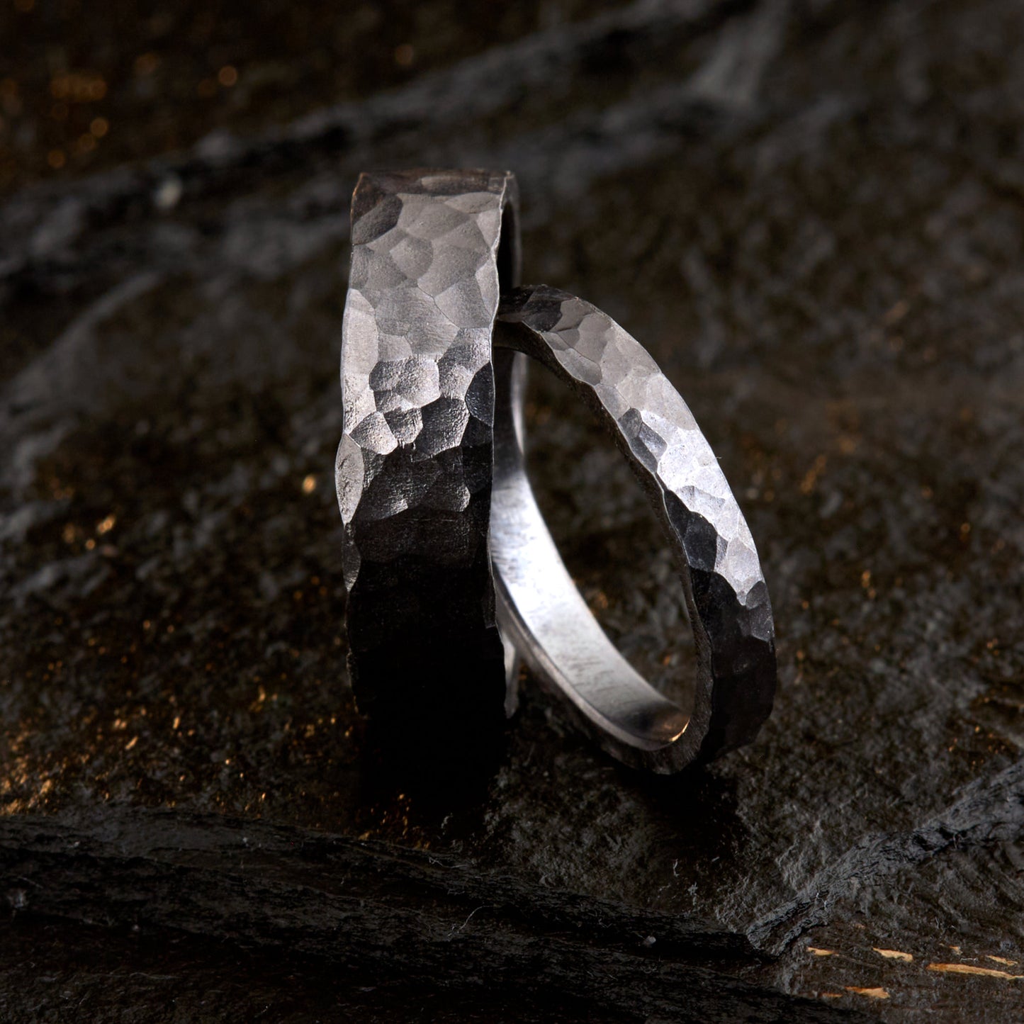 Handgefertigtes Ehering-Set aus massivem Tantal auf rustikalem Leder. Herrenring (6mm) und Damenring (4mm) mit markanter, gehämmerter Oberfläche in dunklem Anthrazit. Einzigartige Schmiedekunst für exklusive Hochzeitsringe. SEO-Keywords: Tantal Eheringe gehämmert, dunkle Trauringe, Tantalum Wedding Bands, nachhaltiger Luxusschmuck, seltene Edelmetalle Hochzeit. Sichtbare Details der individuellen Textur und des tiefen, metallischen Glanzes.