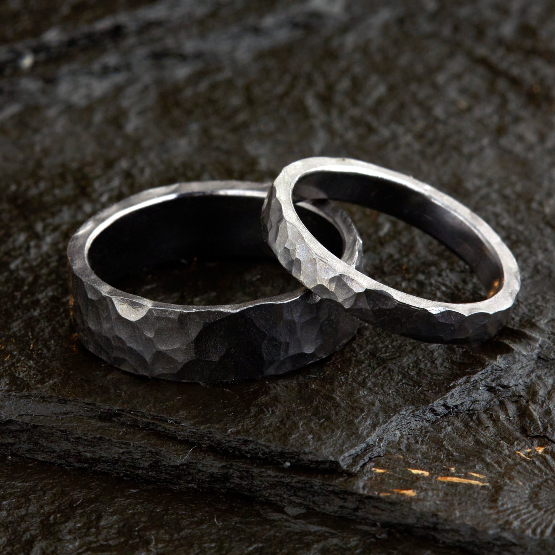 Handgefertigtes Ehering-Set aus massivem Tantal auf rustikalem Leder. Herrenring (6mm) und Damenring (4mm) mit markanter, gehämmerter Oberfläche in dunklem Anthrazit. Einzigartige Schmiedekunst für exklusive Hochzeitsringe. SEO-Keywords: Tantal Eheringe gehämmert, dunkle Trauringe, Tantalum Wedding Bands, nachhaltiger Luxusschmuck, seltene Edelmetalle Hochzeit. Sichtbare Details der individuellen Textur und des tiefen, metallischen Glanzes.