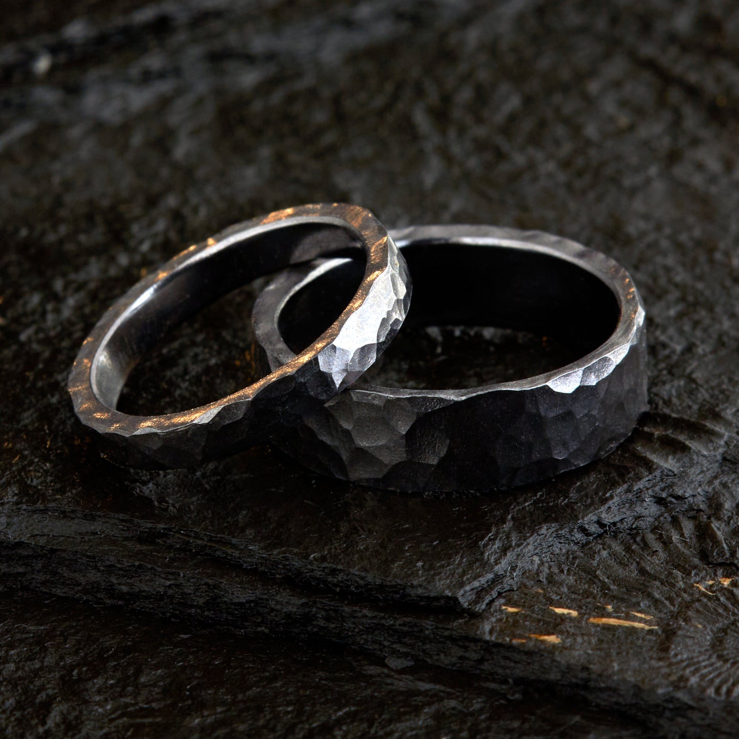 Handgefertigtes Ehering-Set aus massivem Tantal auf rustikalem Leder. Herrenring (6mm) und Damenring (4mm) mit markanter, gehämmerter Oberfläche in dunklem Anthrazit. Einzigartige Schmiedekunst für exklusive Hochzeitsringe. SEO-Keywords: Tantal Eheringe gehämmert, dunkle Trauringe, Tantalum Wedding Bands, nachhaltiger Luxusschmuck, seltene Edelmetalle Hochzeit. Sichtbare Details der individuellen Textur und des tiefen, metallischen Glanzes.