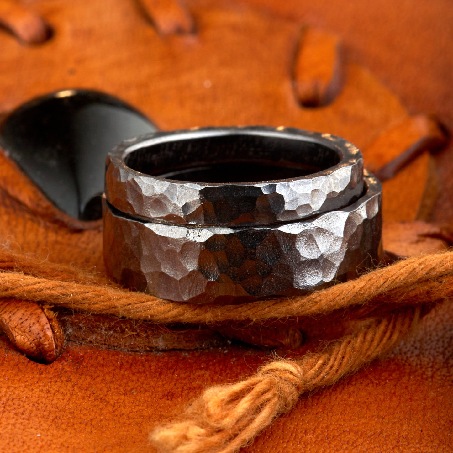 Handgefertigtes Ehering-Set aus massivem Tantal auf rustikalem Leder. Herrenring (6mm) und Damenring (4mm) mit markanter, gehämmerter Oberfläche in dunklem Anthrazit. Einzigartige Schmiedekunst für exklusive Hochzeitsringe. SEO-Keywords: Tantal Eheringe gehämmert, dunkle Trauringe, Tantalum Wedding Bands, nachhaltiger Luxusschmuck, seltene Edelmetalle Hochzeit. Sichtbare Details der individuellen Textur und des tiefen, metallischen Glanzes.