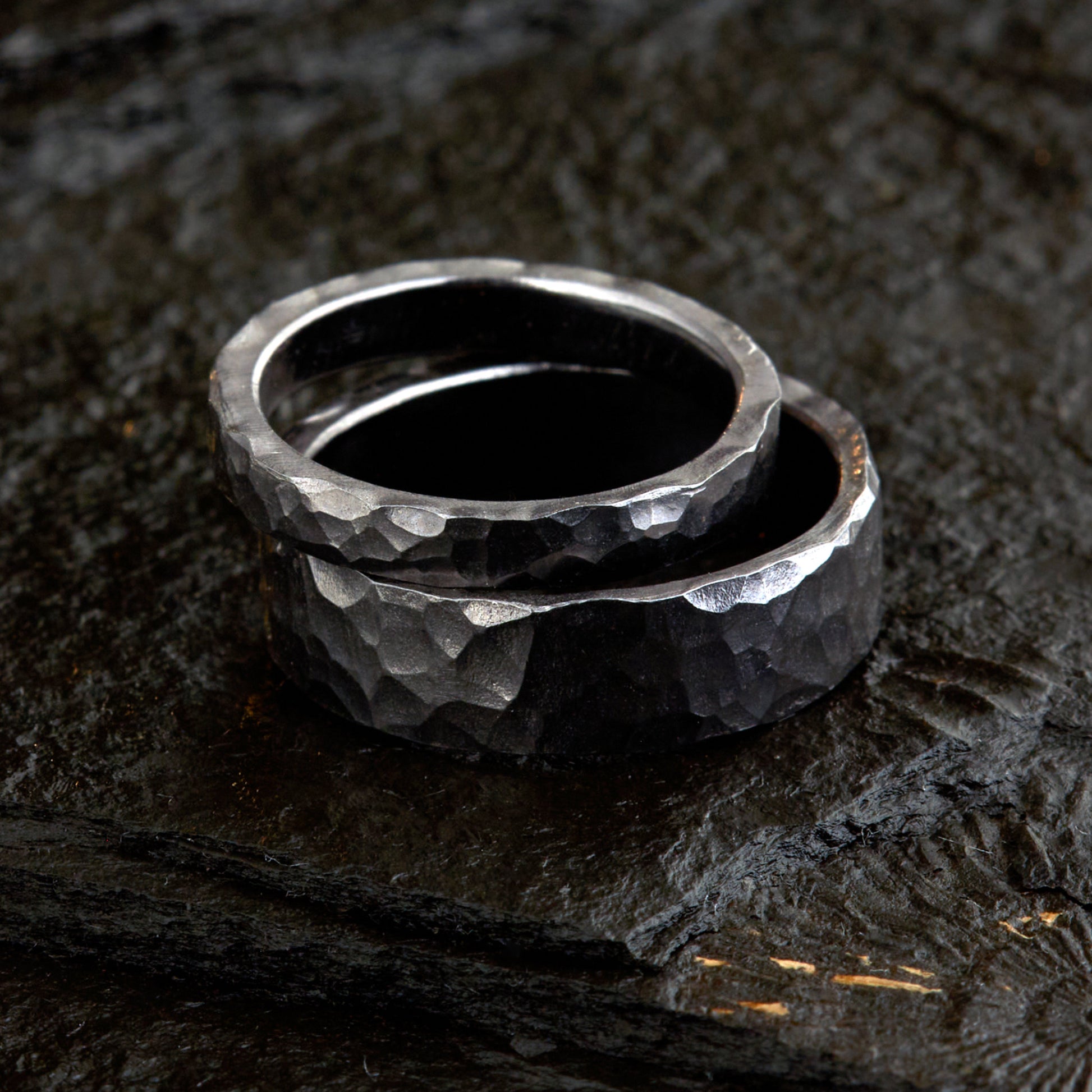 Handgefertigtes Ehering-Set aus massivem Tantal auf rustikalem Leder. Herrenring (6mm) und Damenring (4mm) mit markanter, gehämmerter Oberfläche in dunklem Anthrazit. Einzigartige Schmiedekunst für exklusive Hochzeitsringe. SEO-Keywords: Tantal Eheringe gehämmert, dunkle Trauringe, Tantalum Wedding Bands, nachhaltiger Luxusschmuck, seltene Edelmetalle Hochzeit. Sichtbare Details der individuellen Textur und des tiefen, metallischen Glanzes.