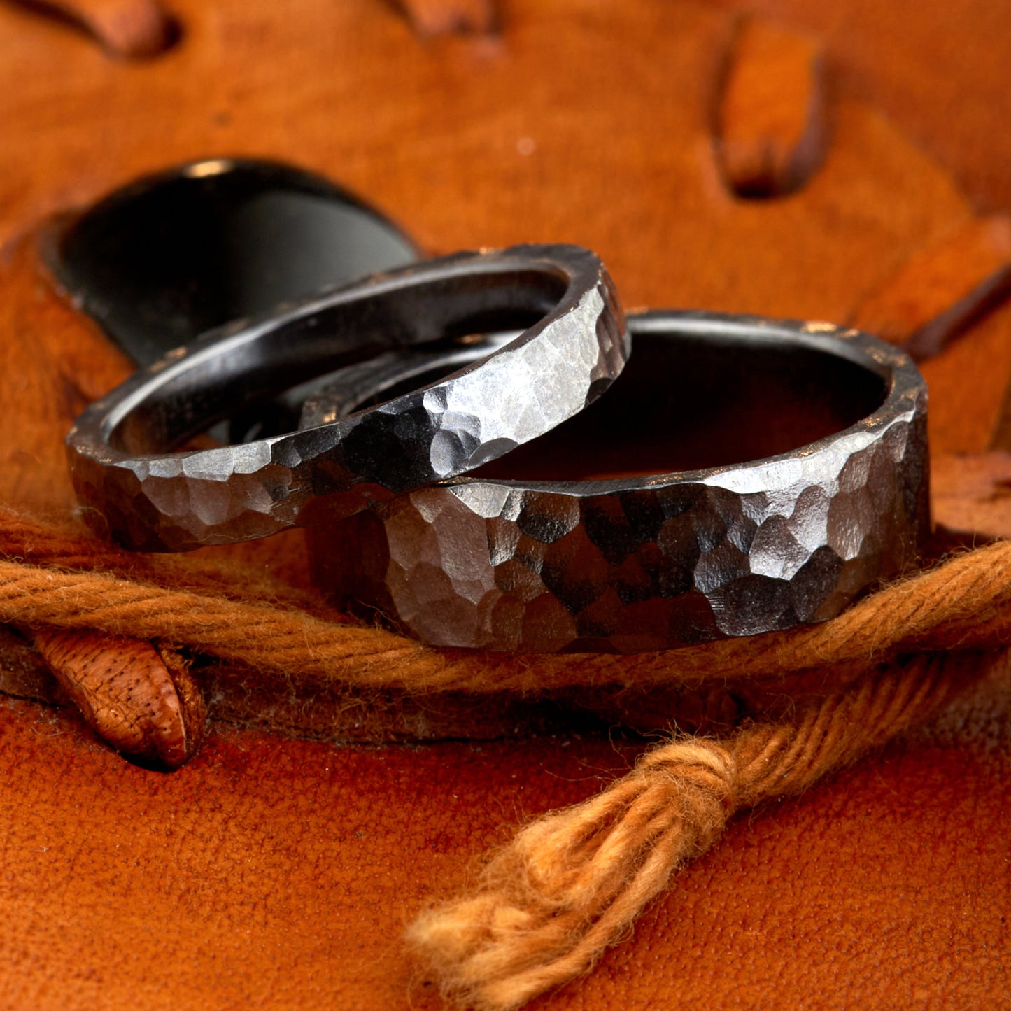 Handgefertigtes Ehering-Set aus massivem Tantal auf rustikalem Leder. Herrenring (6mm) und Damenring (4mm) mit markanter, gehämmerter Oberfläche in dunklem Anthrazit. Einzigartige Schmiedekunst für exklusive Hochzeitsringe. SEO-Keywords: Tantal Eheringe gehämmert, dunkle Trauringe, Tantalum Wedding Bands, nachhaltiger Luxusschmuck, seltene Edelmetalle Hochzeit. Sichtbare Details der individuellen Textur und des tiefen, metallischen Glanzes.