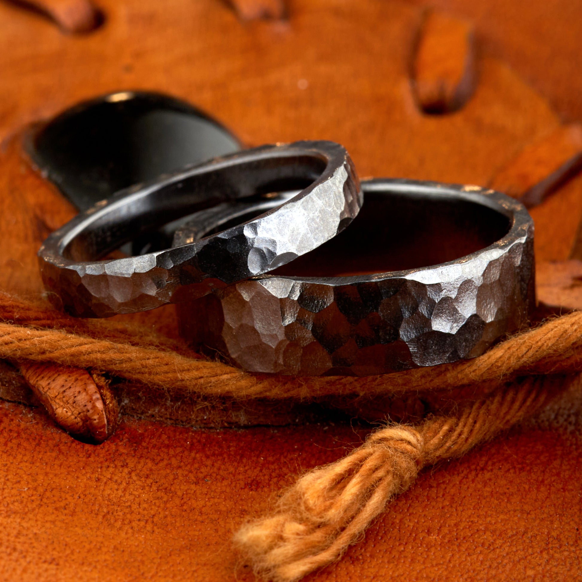 Handgefertigtes Ehering-Set aus massivem Tantal auf rustikalem Leder. Herrenring (6mm) und Damenring (4mm) mit markanter, gehämmerter Oberfläche in dunklem Anthrazit. Einzigartige Schmiedekunst für exklusive Hochzeitsringe. SEO-Keywords: Tantal Eheringe gehämmert, dunkle Trauringe, Tantalum Wedding Bands, nachhaltiger Luxusschmuck, seltene Edelmetalle Hochzeit. Sichtbare Details der individuellen Textur und des tiefen, metallischen Glanzes.