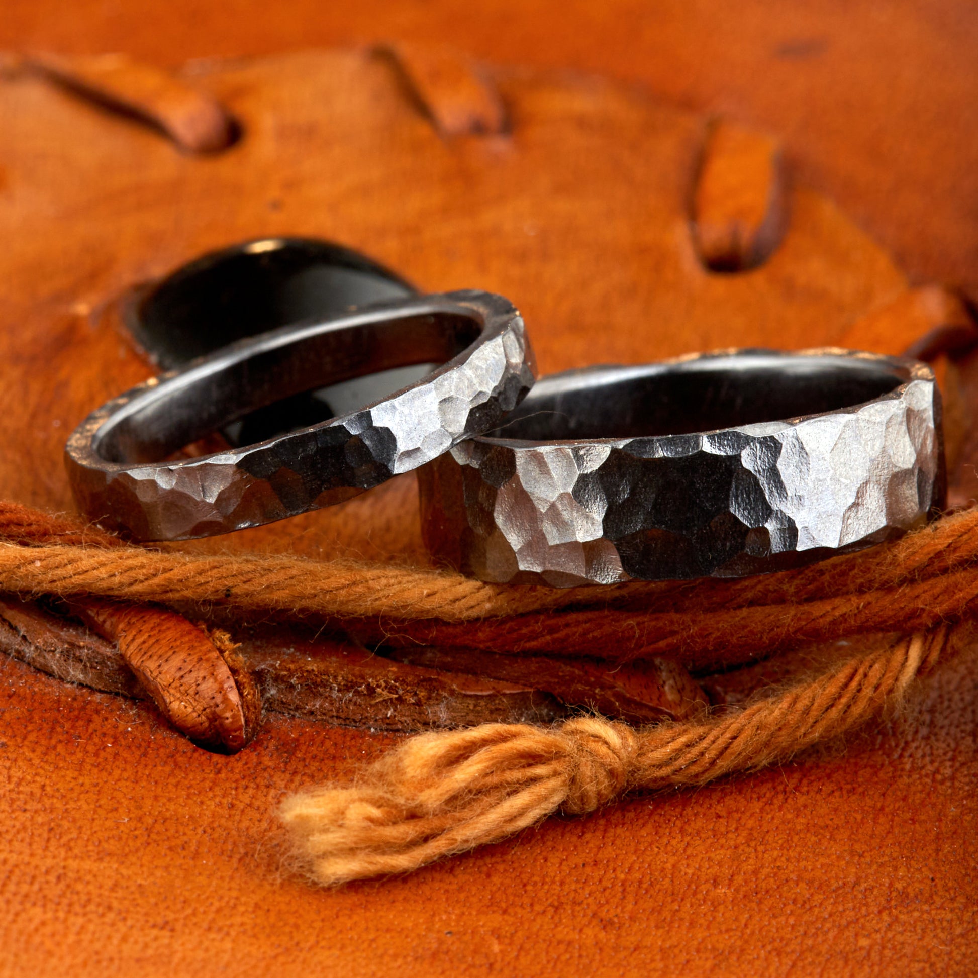 Handgefertigtes Ehering-Set aus massivem Tantal auf rustikalem Leder. Herrenring (6mm) und Damenring (4mm) mit markanter, gehämmerter Oberfläche in dunklem Anthrazit. Einzigartige Schmiedekunst für exklusive Hochzeitsringe. SEO-Keywords: Tantal Eheringe gehämmert, dunkle Trauringe, Tantalum Wedding Bands, nachhaltiger Luxusschmuck, seltene Edelmetalle Hochzeit. Sichtbare Details der individuellen Textur und des tiefen, metallischen Glanzes.