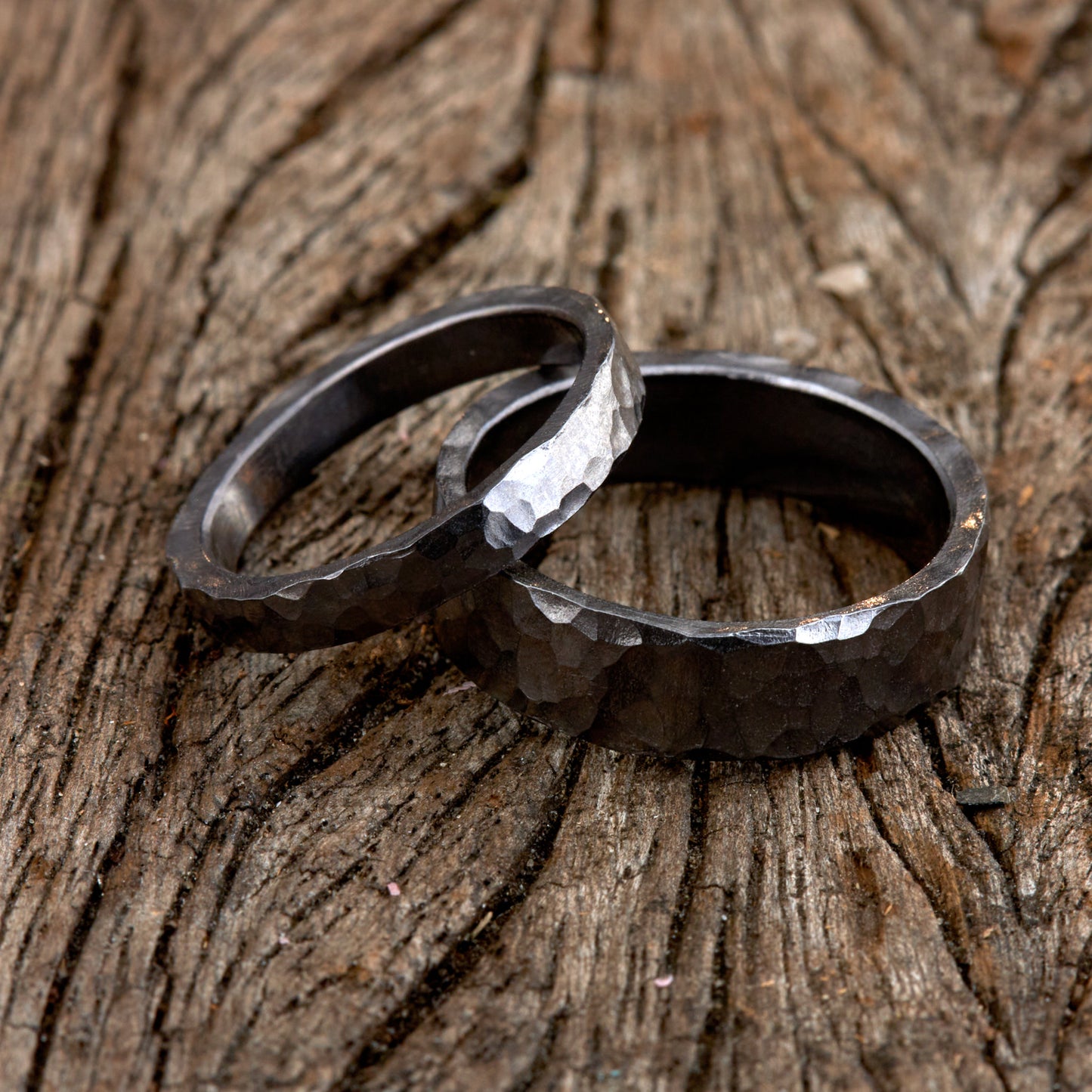 Handgefertigtes Ehering-Set aus massivem Tantal auf rustikalem Leder. Herrenring (6mm) und Damenring (4mm) mit markanter, gehämmerter Oberfläche in dunklem Anthrazit. Einzigartige Schmiedekunst für exklusive Hochzeitsringe. SEO-Keywords: Tantal Eheringe gehämmert, dunkle Trauringe, Tantalum Wedding Bands, nachhaltiger Luxusschmuck, seltene Edelmetalle Hochzeit. Sichtbare Details der individuellen Textur und des tiefen, metallischen Glanzes.