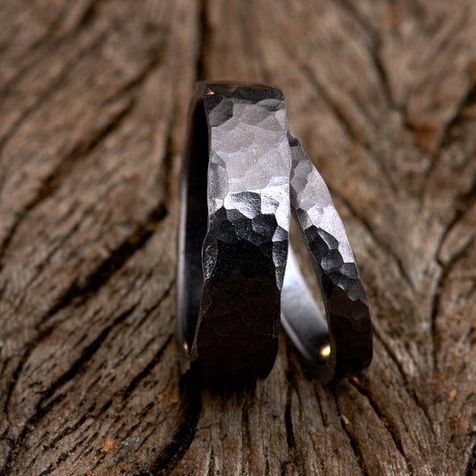 Handgefertigtes Ehering-Set aus massivem Tantal auf rustikalem Leder. Herrenring (6mm) und Damenring (4mm) mit markanter, gehämmerter Oberfläche in dunklem Anthrazit. Einzigartige Schmiedekunst für exklusive Hochzeitsringe. SEO-Keywords: Tantal Eheringe gehämmert, dunkle Trauringe, Tantalum Wedding Bands, nachhaltiger Luxusschmuck, seltene Edelmetalle Hochzeit. Sichtbare Details der individuellen Textur und des tiefen, metallischen Glanzes.