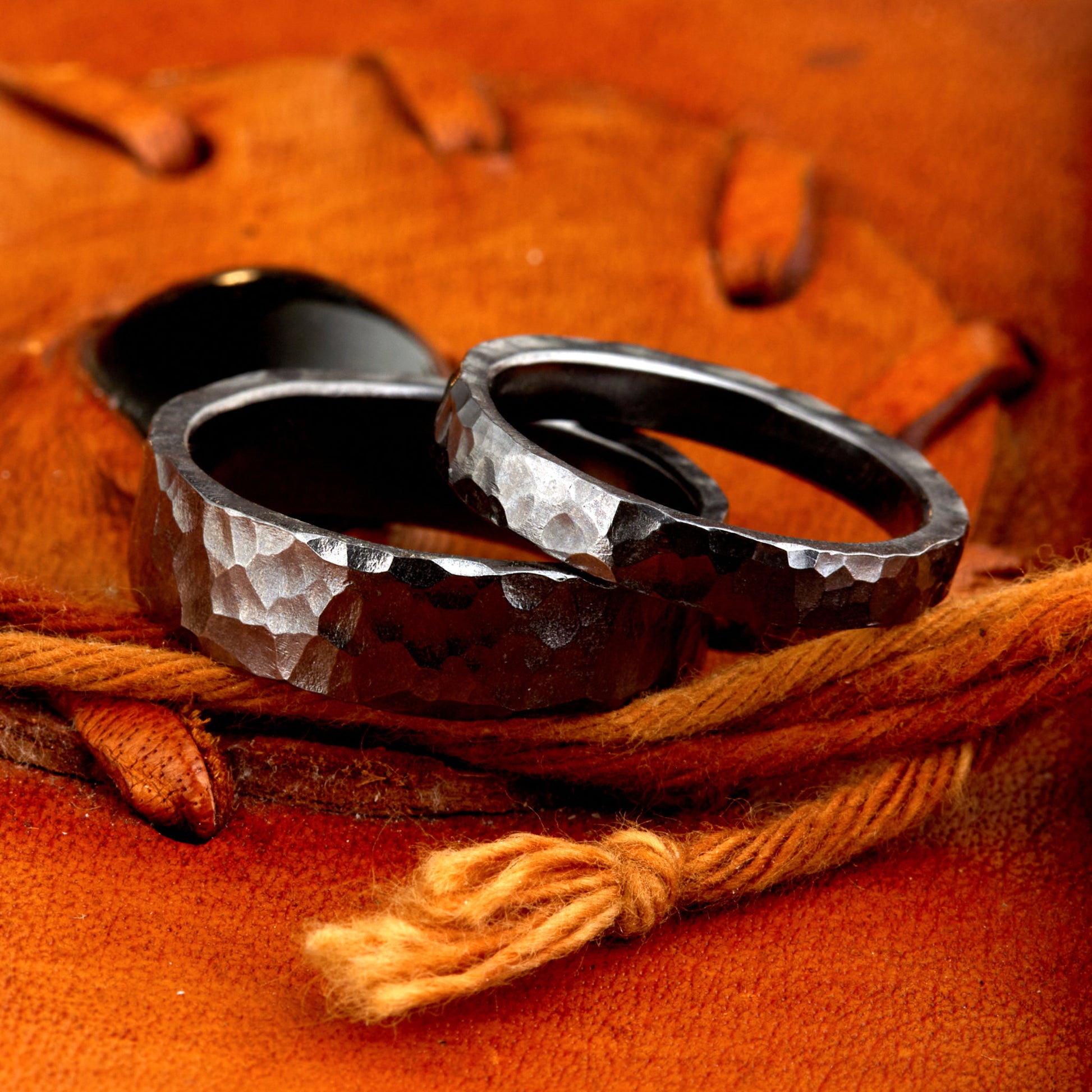 Handgefertigtes Ehering-Set aus massivem Tantal auf rustikalem Leder. Herrenring (6mm) und Damenring (4mm) mit markanter, gehämmerter Oberfläche in dunklem Anthrazit. Einzigartige Schmiedekunst für exklusive Hochzeitsringe. SEO-Keywords: Tantal Eheringe gehämmert, dunkle Trauringe, Tantalum Wedding Bands, nachhaltiger Luxusschmuck, seltene Edelmetalle Hochzeit. Sichtbare Details der individuellen Textur und des tiefen, metallischen Glanzes.