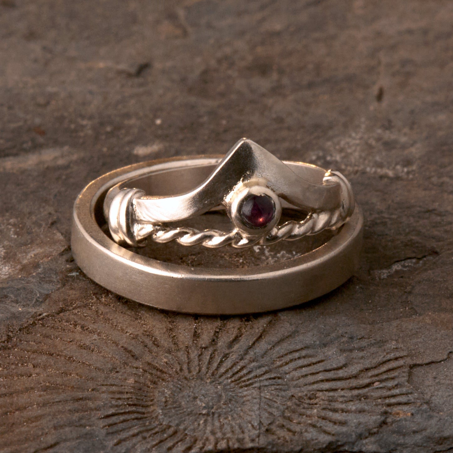 Handgefertigter Vintage Wishbone-Ring aus mattem 925 Silber mit einem violetten 3 mm Amethyst Cabochon in Zargenfassung. Man sieht deutlich die geteilte Schiene mit der filigranen Kordel-Verblendung.
Set-Präsentation: Minimalistisches Ehering-Set von Silberschweif auf dunklem Stein. Der verspielte Damenring mit lila Edelstein harmoniert perfekt mit dem schlichten, 4 mm breiten matten Herrenring.
Detail Ringschiene: Seitenansicht des Damenrings zeigt die handwerkliche Aufteilung der 4 mm Silberschiene 
