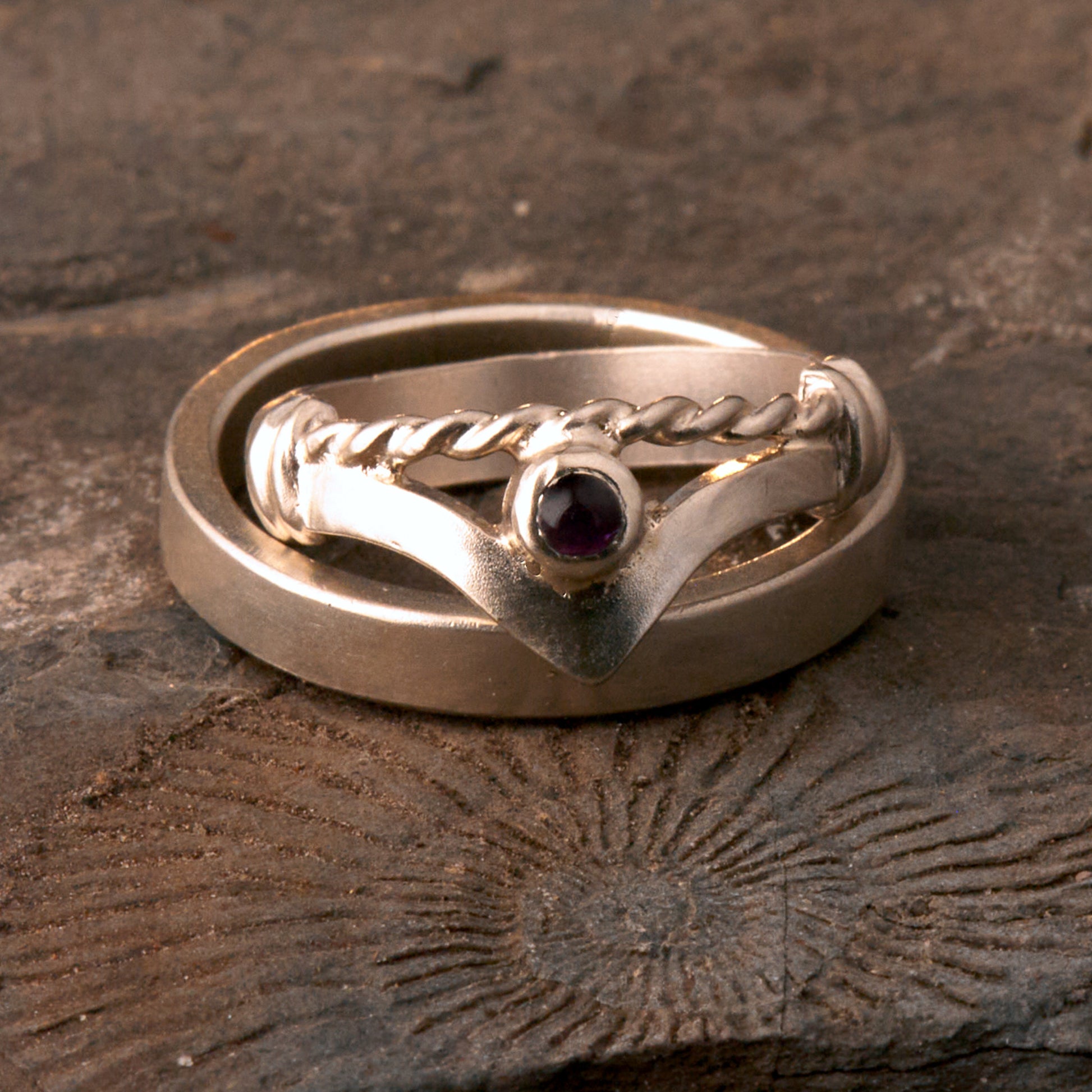 Handgefertigter Vintage Wishbone-Ring aus mattem 925 Silber mit einem violetten 3 mm Amethyst Cabochon in Zargenfassung. Man sieht deutlich die geteilte Schiene mit der filigranen Kordel-Verblendung.
Set-Präsentation: Minimalistisches Ehering-Set von Silberschweif auf dunklem Stein. Der verspielte Damenring mit lila Edelstein harmoniert perfekt mit dem schlichten, 4 mm breiten matten Herrenring.
Detail Ringschiene: Seitenansicht des Damenrings zeigt die handwerkliche Aufteilung der 4 mm Silberschiene 