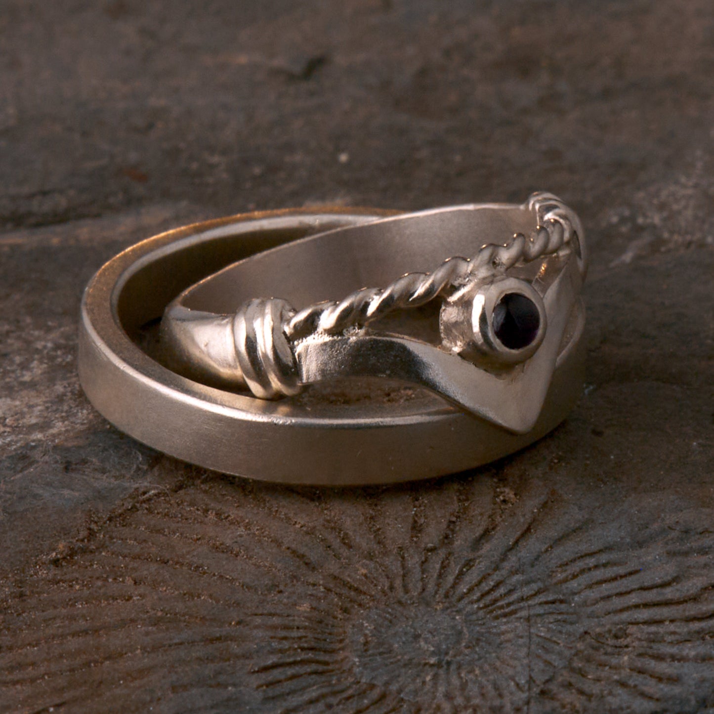 Handgefertigter Vintage Wishbone-Ring aus mattem 925 Silber mit einem violetten 3 mm Amethyst Cabochon in Zargenfassung. Man sieht deutlich die geteilte Schiene mit der filigranen Kordel-Verblendung.
Set-Präsentation: Minimalistisches Ehering-Set von Silberschweif auf dunklem Stein. Der verspielte Damenring mit lila Edelstein harmoniert perfekt mit dem schlichten, 4 mm breiten matten Herrenring.
Detail Ringschiene: Seitenansicht des Damenrings zeigt die handwerkliche Aufteilung der 4 mm Silberschiene 