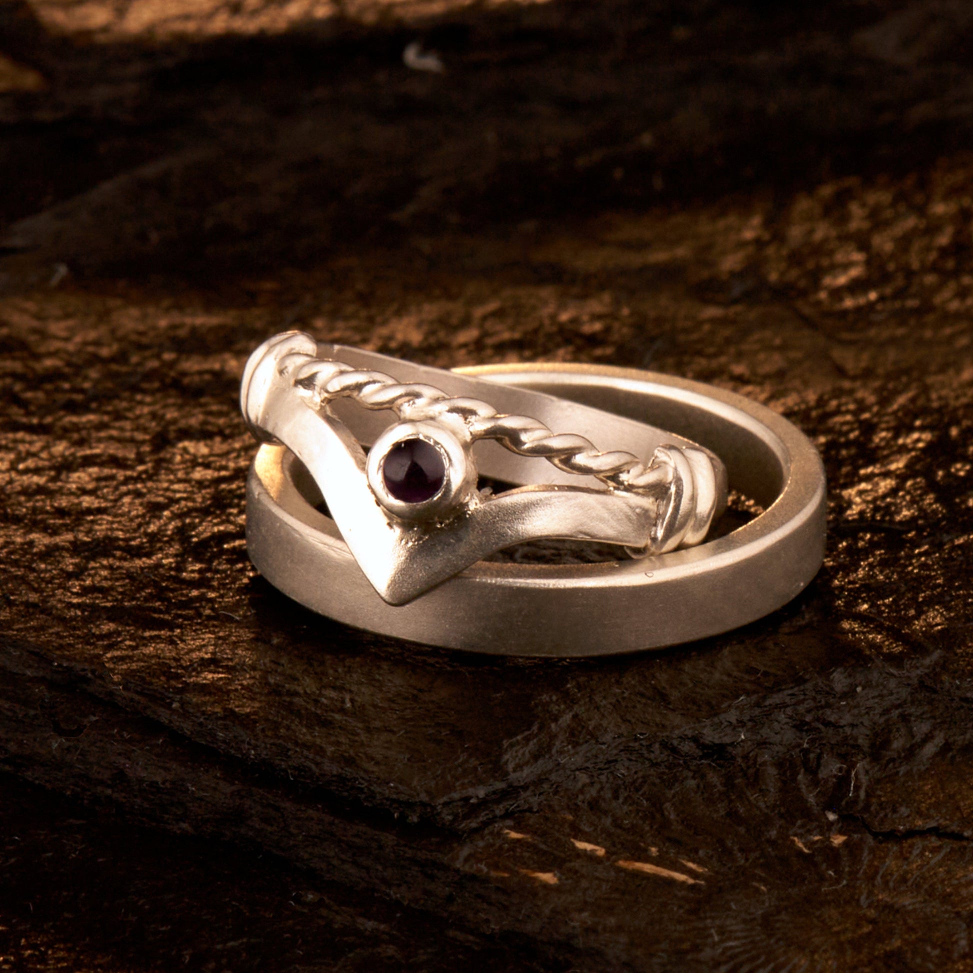 Handgefertigter Vintage Wishbone-Ring aus mattem 925 Silber mit einem violetten 3 mm Amethyst Cabochon in Zargenfassung. Man sieht deutlich die geteilte Schiene mit der filigranen Kordel-Verblendung.
Set-Präsentation: Minimalistisches Ehering-Set von Silberschweif auf dunklem Stein. Der verspielte Damenring mit lila Edelstein harmoniert perfekt mit dem schlichten, 4 mm breiten matten Herrenring.
Detail Ringschiene: Seitenansicht des Damenrings zeigt die handwerkliche Aufteilung der 4 mm Silberschiene 