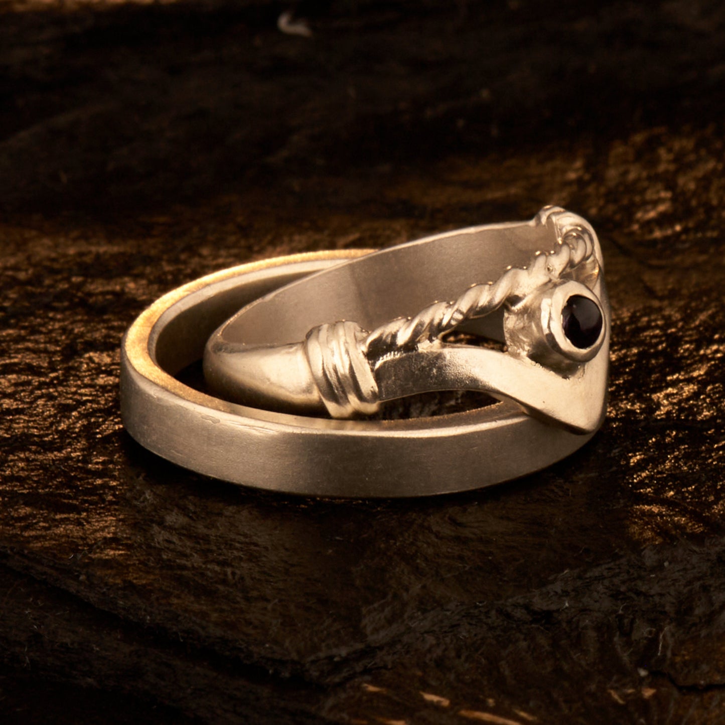 Handgefertigter Vintage Wishbone-Ring aus mattem 925 Silber mit einem violetten 3 mm Amethyst Cabochon in Zargenfassung. Man sieht deutlich die geteilte Schiene mit der filigranen Kordel-Verblendung.
Set-Präsentation: Minimalistisches Ehering-Set von Silberschweif auf dunklem Stein. Der verspielte Damenring mit lila Edelstein harmoniert perfekt mit dem schlichten, 4 mm breiten matten Herrenring.
Detail Ringschiene: Seitenansicht des Damenrings zeigt die handwerkliche Aufteilung der 4 mm Silberschiene 