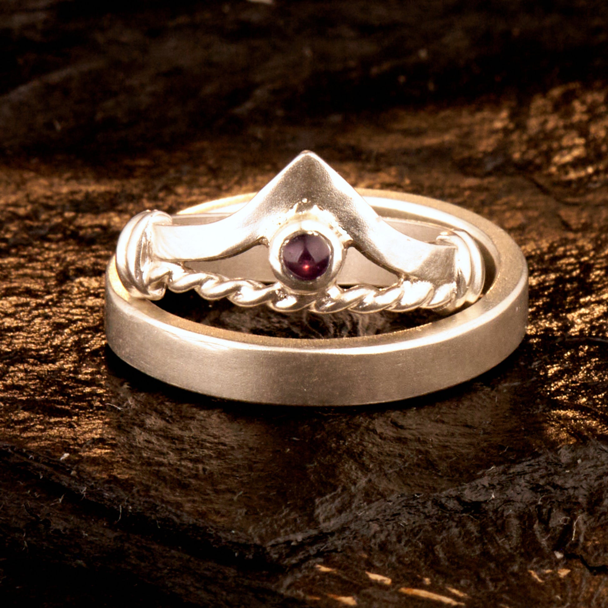 Handgefertigter Vintage Wishbone-Ring aus mattem 925 Silber mit einem violetten 3 mm Amethyst Cabochon in Zargenfassung. Man sieht deutlich die geteilte Schiene mit der filigranen Kordel-Verblendung.
Set-Präsentation: Minimalistisches Ehering-Set von Silberschweif auf dunklem Stein. Der verspielte Damenring mit lila Edelstein harmoniert perfekt mit dem schlichten, 4 mm breiten matten Herrenring.
Detail Ringschiene: Seitenansicht des Damenrings zeigt die handwerkliche Aufteilung der 4 mm Silberschiene 
