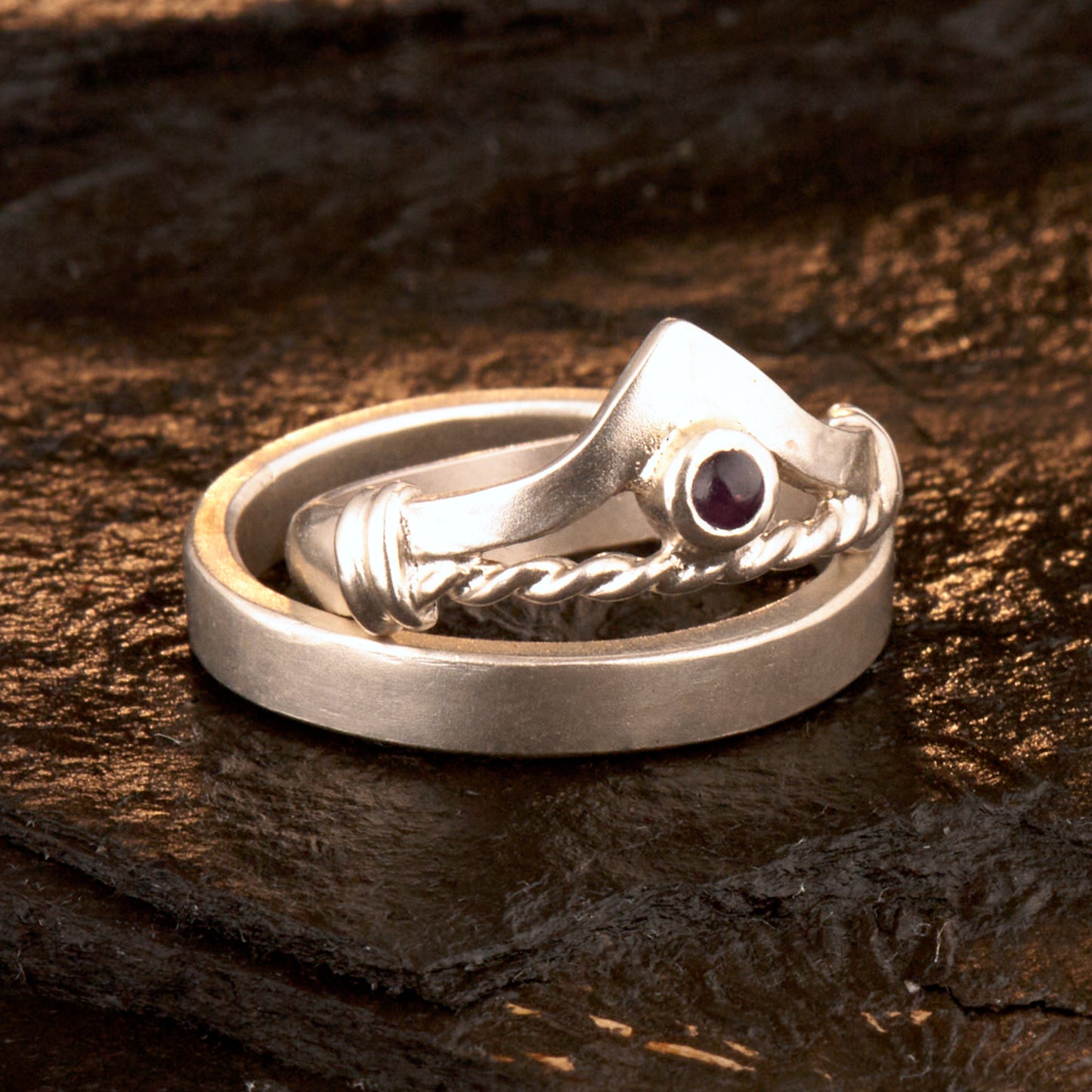 Handgefertigter Vintage Wishbone-Ring aus mattem 925 Silber mit einem violetten 3 mm Amethyst Cabochon in Zargenfassung. Man sieht deutlich die geteilte Schiene mit der filigranen Kordel-Verblendung.
Set-Präsentation: Minimalistisches Ehering-Set von Silberschweif auf dunklem Stein. Der verspielte Damenring mit lila Edelstein harmoniert perfekt mit dem schlichten, 4 mm breiten matten Herrenring.
Detail Ringschiene: Seitenansicht des Damenrings zeigt die handwerkliche Aufteilung der 4 mm Silberschiene 