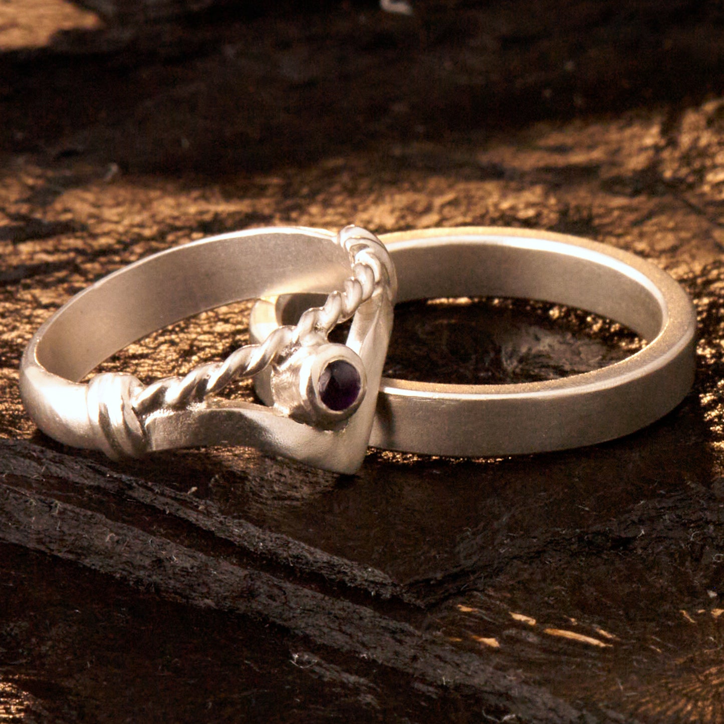 Handgefertigter Vintage Wishbone-Ring aus mattem 925 Silber mit einem violetten 3 mm Amethyst Cabochon in Zargenfassung. Man sieht deutlich die geteilte Schiene mit der filigranen Kordel-Verblendung.
Set-Präsentation: Minimalistisches Ehering-Set von Silberschweif auf dunklem Stein. Der verspielte Damenring mit lila Edelstein harmoniert perfekt mit dem schlichten, 4 mm breiten matten Herrenring.
Detail Ringschiene: Seitenansicht des Damenrings zeigt die handwerkliche Aufteilung der 4 mm Silberschiene 
