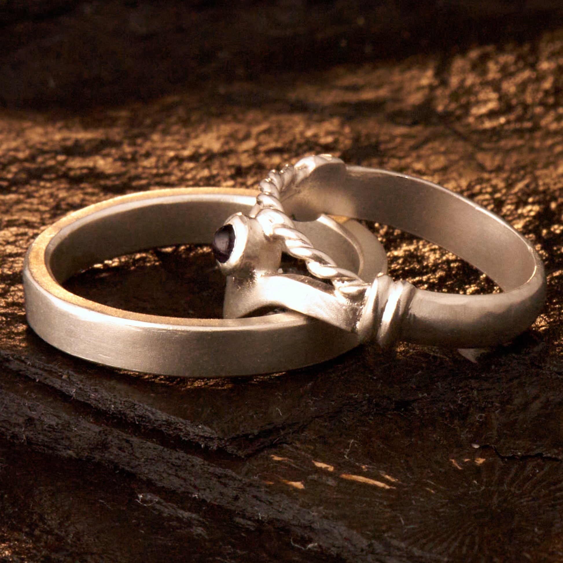 Handgefertigter Vintage Wishbone-Ring aus mattem 925 Silber mit einem violetten 3 mm Amethyst Cabochon in Zargenfassung. Man sieht deutlich die geteilte Schiene mit der filigranen Kordel-Verblendung.
Set-Präsentation: Minimalistisches Ehering-Set von Silberschweif auf dunklem Stein. Der verspielte Damenring mit lila Edelstein harmoniert perfekt mit dem schlichten, 4 mm breiten matten Herrenring.
Detail Ringschiene: Seitenansicht des Damenrings zeigt die handwerkliche Aufteilung der 4 mm Silberschiene 
