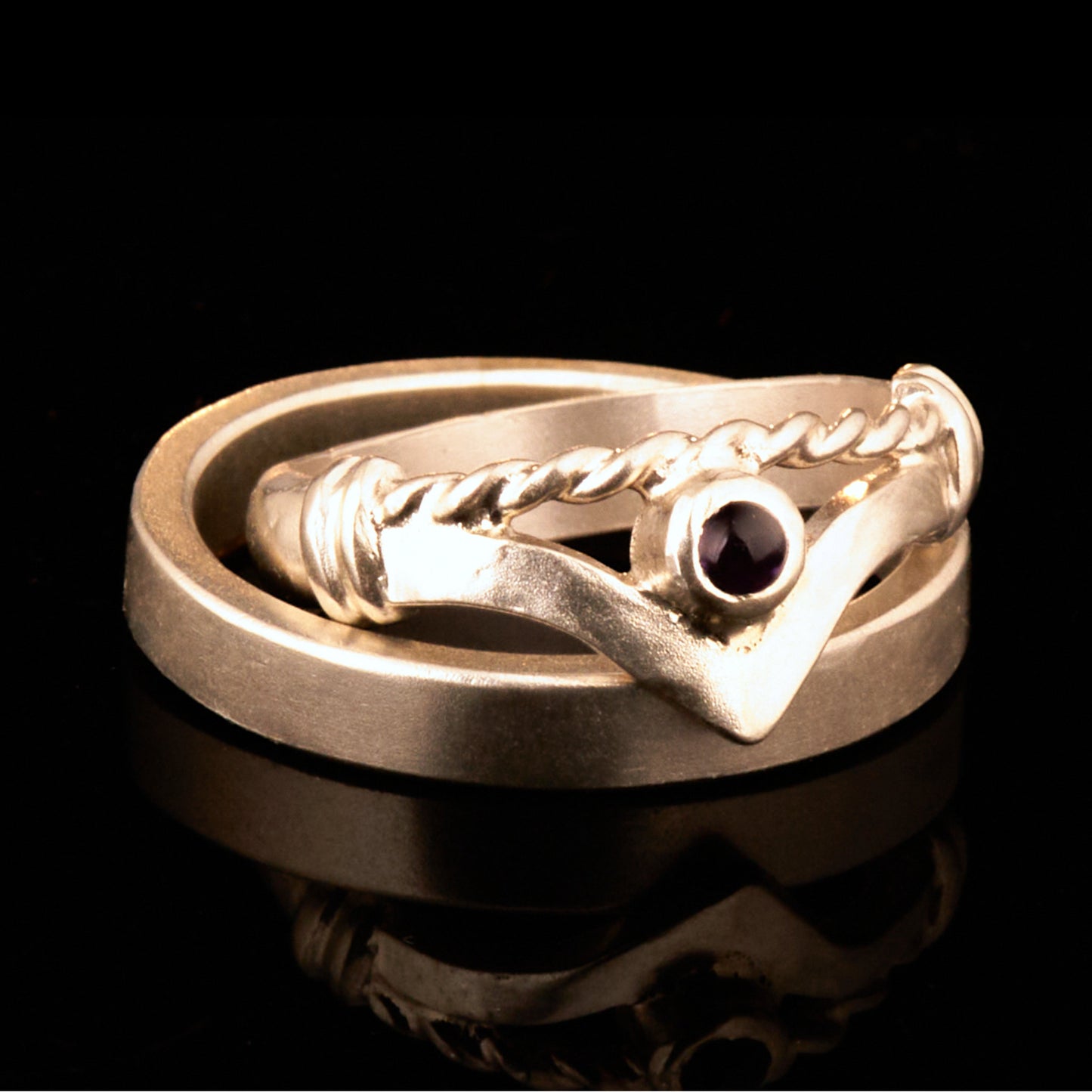 Handgefertigter Vintage Wishbone-Ring aus mattem 925 Silber mit einem violetten 3 mm Amethyst Cabochon in Zargenfassung. Man sieht deutlich die geteilte Schiene mit der filigranen Kordel-Verblendung.
Set-Präsentation: Minimalistisches Ehering-Set von Silberschweif auf dunklem Stein. Der verspielte Damenring mit lila Edelstein harmoniert perfekt mit dem schlichten, 4 mm breiten matten Herrenring.
Detail Ringschiene: Seitenansicht des Damenrings zeigt die handwerkliche Aufteilung der 4 mm Silberschiene 