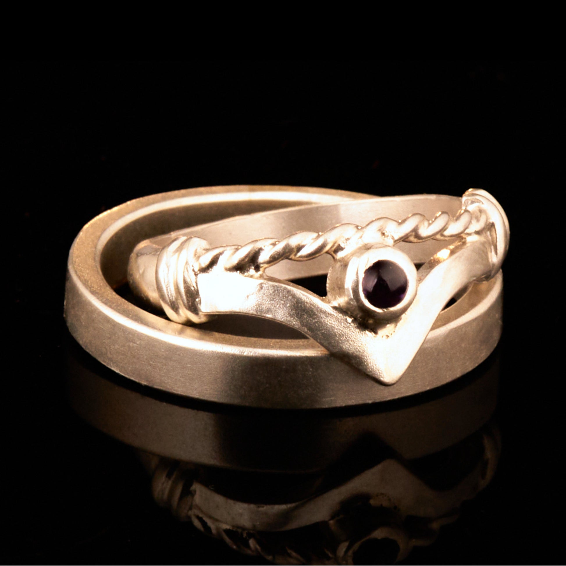 Handgefertigter Vintage Wishbone-Ring aus mattem 925 Silber mit einem violetten 3 mm Amethyst Cabochon in Zargenfassung. Man sieht deutlich die geteilte Schiene mit der filigranen Kordel-Verblendung.
Set-Präsentation: Minimalistisches Ehering-Set von Silberschweif auf dunklem Stein. Der verspielte Damenring mit lila Edelstein harmoniert perfekt mit dem schlichten, 4 mm breiten matten Herrenring.
Detail Ringschiene: Seitenansicht des Damenrings zeigt die handwerkliche Aufteilung der 4 mm Silberschiene 