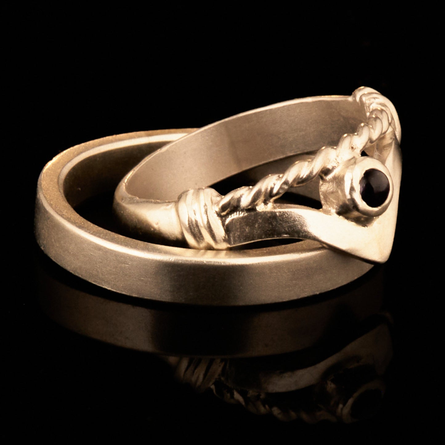 Handgefertigter Vintage Wishbone-Ring aus mattem 925 Silber mit einem violetten 3 mm Amethyst Cabochon in Zargenfassung. Man sieht deutlich die geteilte Schiene mit der filigranen Kordel-Verblendung.
Set-Präsentation: Minimalistisches Ehering-Set von Silberschweif auf dunklem Stein. Der verspielte Damenring mit lila Edelstein harmoniert perfekt mit dem schlichten, 4 mm breiten matten Herrenring.
Detail Ringschiene: Seitenansicht des Damenrings zeigt die handwerkliche Aufteilung der 4 mm Silberschiene 