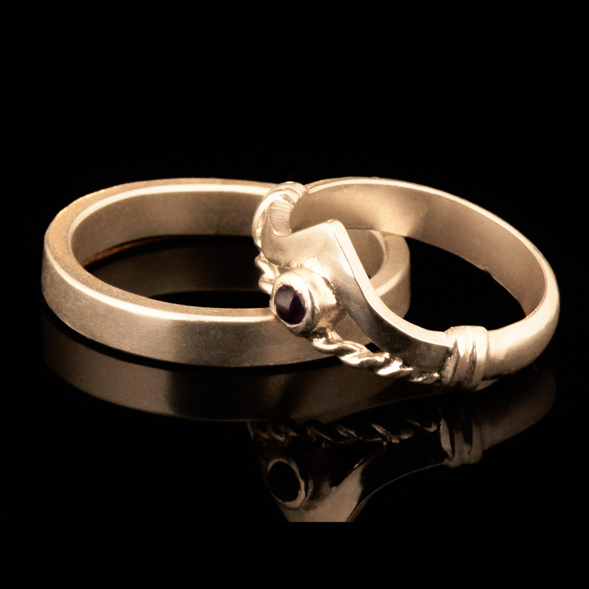 Handgefertigter Vintage Wishbone-Ring aus mattem 925 Silber mit einem violetten 3 mm Amethyst Cabochon in Zargenfassung. Man sieht deutlich die geteilte Schiene mit der filigranen Kordel-Verblendung.
Set-Präsentation: Minimalistisches Ehering-Set von Silberschweif auf dunklem Stein. Der verspielte Damenring mit lila Edelstein harmoniert perfekt mit dem schlichten, 4 mm breiten matten Herrenring.
Detail Ringschiene: Seitenansicht des Damenrings zeigt die handwerkliche Aufteilung der 4 mm Silberschiene 