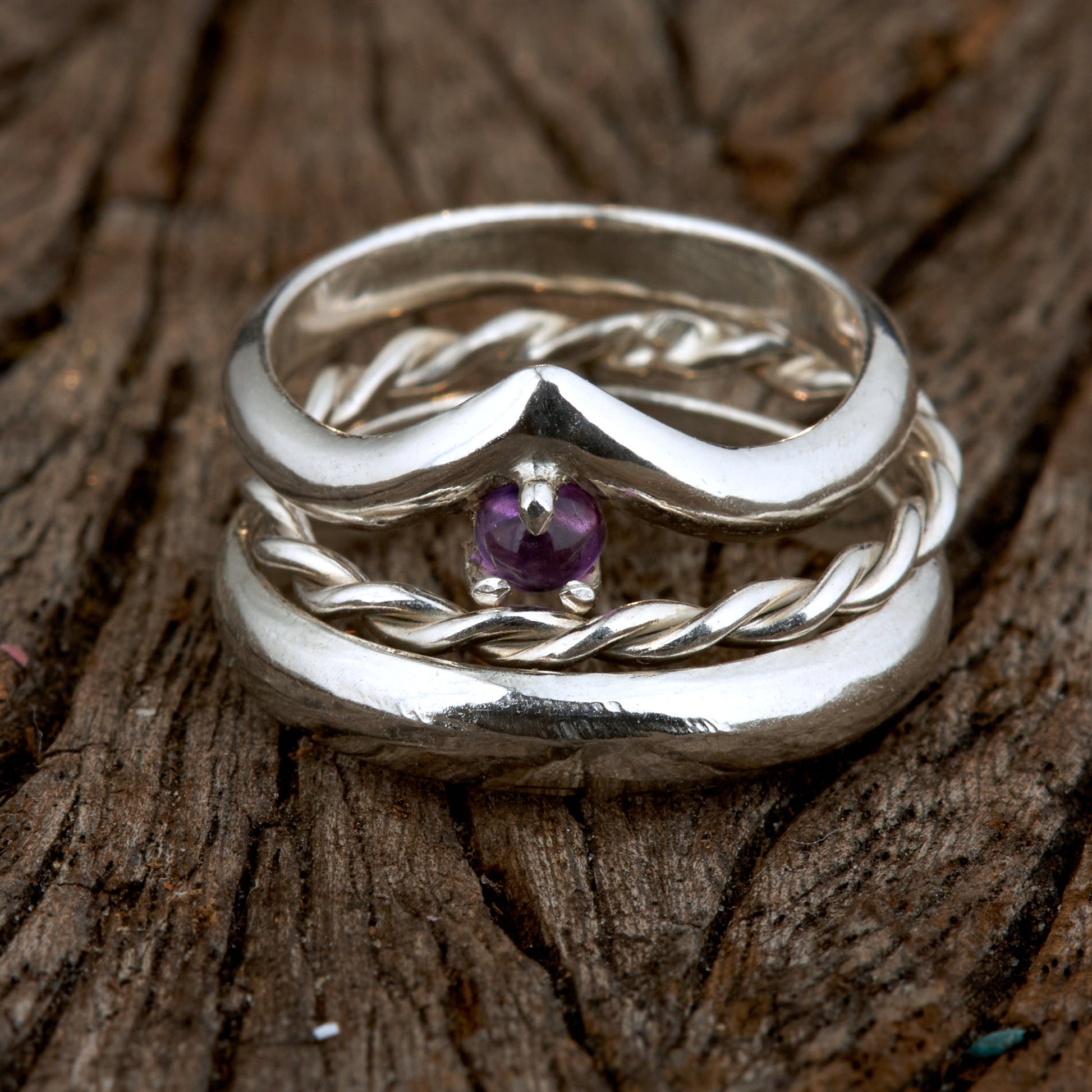 Handgefertigtes Ehering-Set aus 925 Silber auf Leder. 3-teiliges Damen-Set bestehend aus einem 3mm Wishbone-Ring mit Amethyst und einem 2mm Kordelring. Daneben ein massiver 4mm Herrenring im Halbrund-Profil. Zeitloses Design mit lila Edelstein für die Hochzeit. SEO-Keywords: Wishbone Eheringe Silber, Amethyst Trauringe Set, handgefertigte Silberringe, 925 Sterlingsilber Schmuck, Kordelring Hochzeit. Sichtbare Details der hochwertigen Handarbeit und Steinbesetzung.