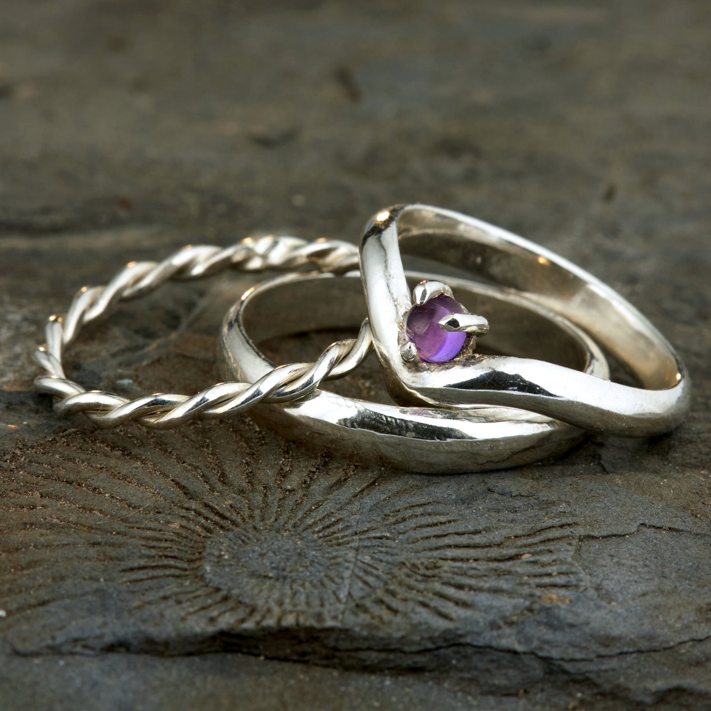 Handgefertigtes Ehering-Set aus 925 Silber auf Leder. 3-teiliges Damen-Set bestehend aus einem 3mm Wishbone-Ring mit Amethyst und einem 2mm Kordelring. Daneben ein massiver 4mm Herrenring im Halbrund-Profil. Zeitloses Design mit lila Edelstein für die Hochzeit. SEO-Keywords: Wishbone Eheringe Silber, Amethyst Trauringe Set, handgefertigte Silberringe, 925 Sterlingsilber Schmuck, Kordelring Hochzeit. Sichtbare Details der hochwertigen Handarbeit und Steinbesetzung.
