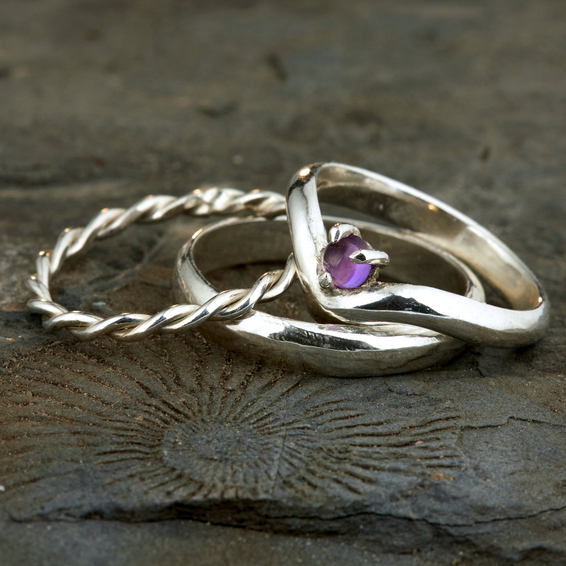 Handgefertigtes Ehering-Set aus 925 Silber auf Leder. 3-teiliges Damen-Set bestehend aus einem 3mm Wishbone-Ring mit Amethyst und einem 2mm Kordelring. Daneben ein massiver 4mm Herrenring im Halbrund-Profil. Zeitloses Design mit lila Edelstein für die Hochzeit. SEO-Keywords: Wishbone Eheringe Silber, Amethyst Trauringe Set, handgefertigte Silberringe, 925 Sterlingsilber Schmuck, Kordelring Hochzeit. Sichtbare Details der hochwertigen Handarbeit und Steinbesetzung.