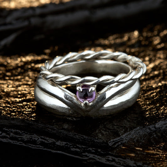 Handgefertigtes Ehering-Set aus 925 Silber auf Leder. 3-teiliges Damen-Set bestehend aus einem 3mm Wishbone-Ring mit Amethyst und einem 2mm Kordelring. Daneben ein massiver 4mm Herrenring im Halbrund-Profil. Zeitloses Design mit lila Edelstein für die Hochzeit. SEO-Keywords: Wishbone Eheringe Silber, Amethyst Trauringe Set, handgefertigte Silberringe, 925 Sterlingsilber Schmuck, Kordelring Hochzeit. Sichtbare Details der hochwertigen Handarbeit und Steinbesetzung.