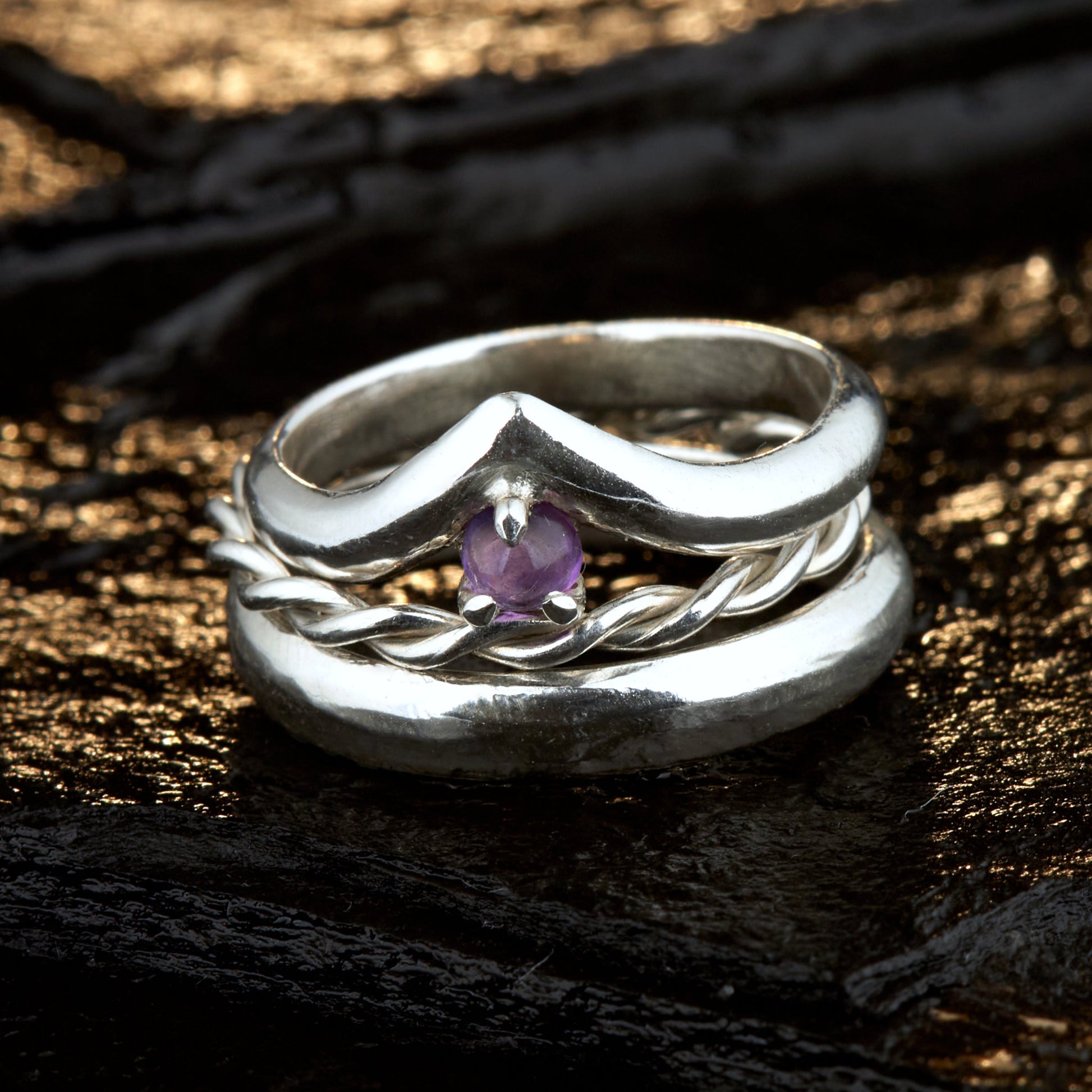 Handgefertigtes Ehering-Set aus 925 Silber auf Leder. 3-teiliges Damen-Set bestehend aus einem 3mm Wishbone-Ring mit Amethyst und einem 2mm Kordelring. Daneben ein massiver 4mm Herrenring im Halbrund-Profil. Zeitloses Design mit lila Edelstein für die Hochzeit. SEO-Keywords: Wishbone Eheringe Silber, Amethyst Trauringe Set, handgefertigte Silberringe, 925 Sterlingsilber Schmuck, Kordelring Hochzeit. Sichtbare Details der hochwertigen Handarbeit und Steinbesetzung.